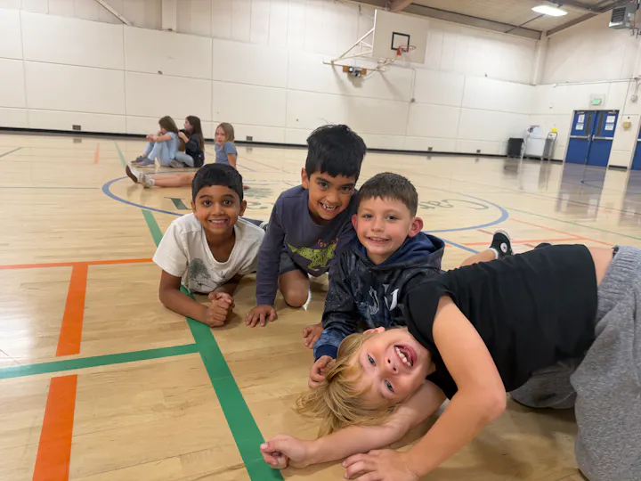 Four boys are playfully lying on the gym floor, smiling at the camera, while some girls sit in the background.