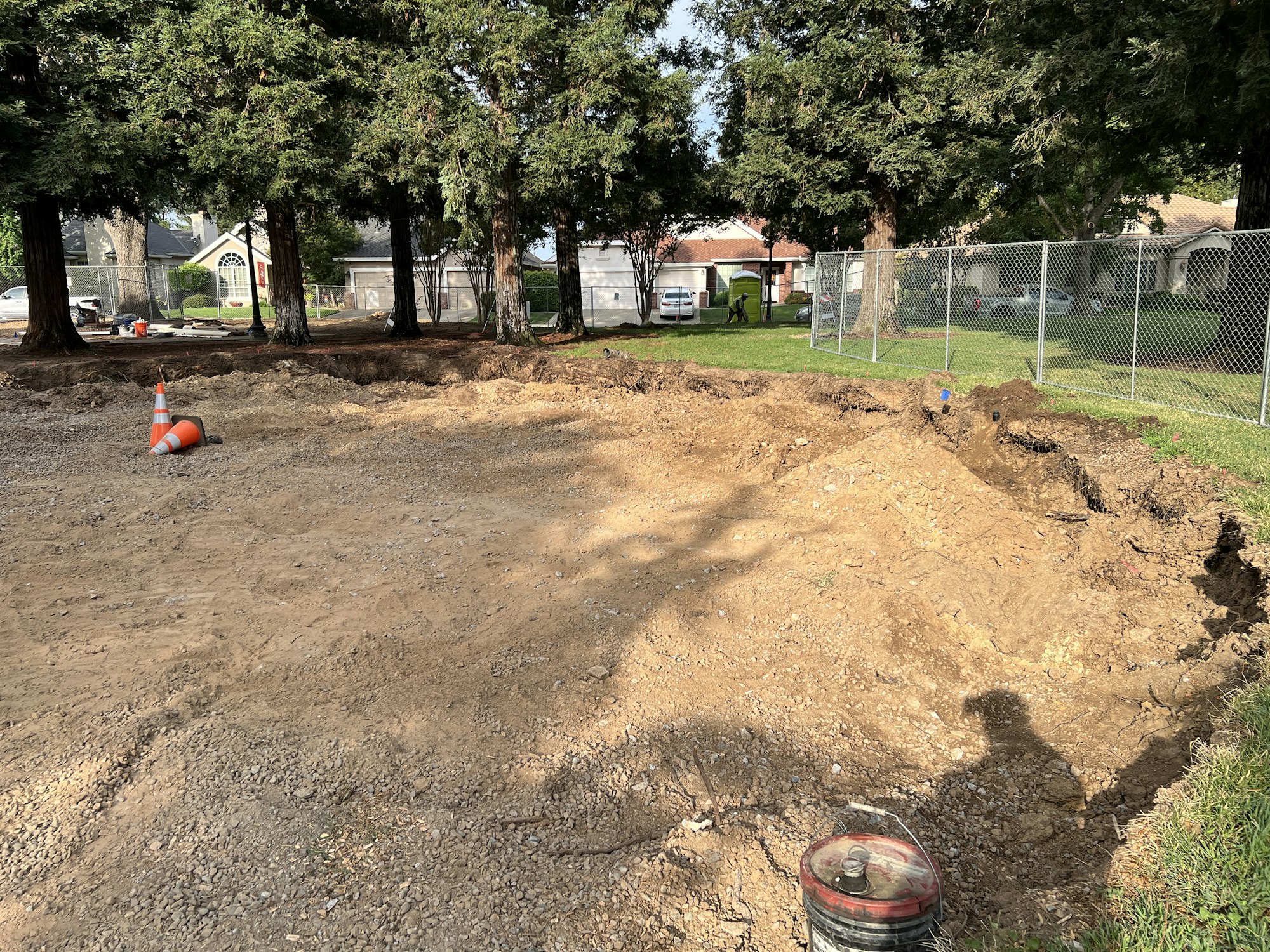 A construction site with dirt, orange cones, a fence, trees, and nearby houses.