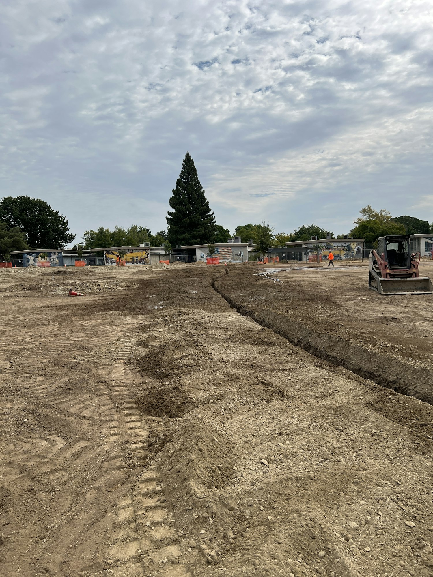 Construction site with dug-up ground, machinery, and a person in an orange vest. Cloudy sky and trees in the background.