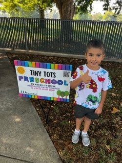 A child standing next to a "Tiny Tots Preschool" sign, smiling and giving a thumbs-up.