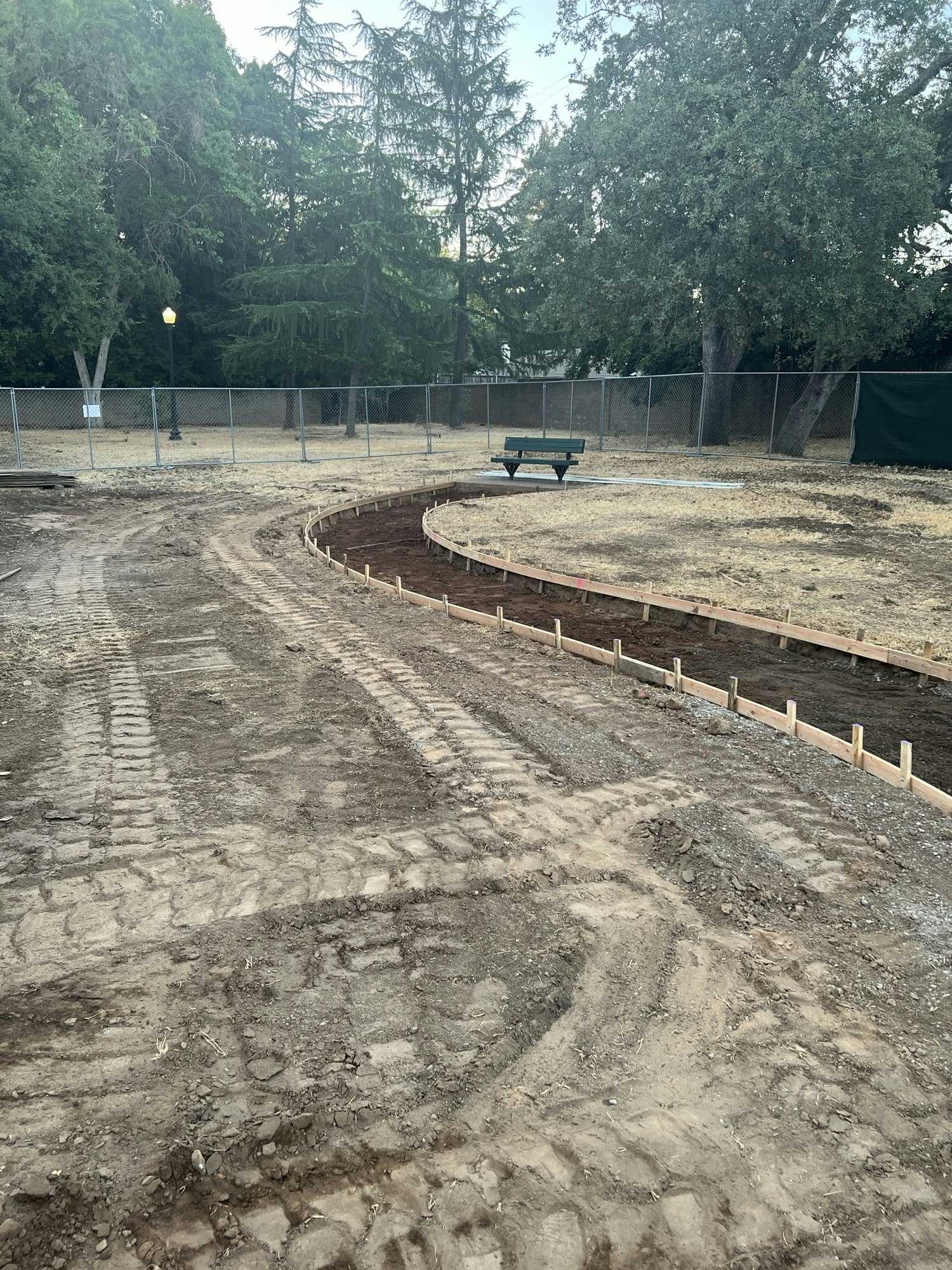 A construction site with dirt paths, a curved area, wooden forms, a bench, and trees in the background.