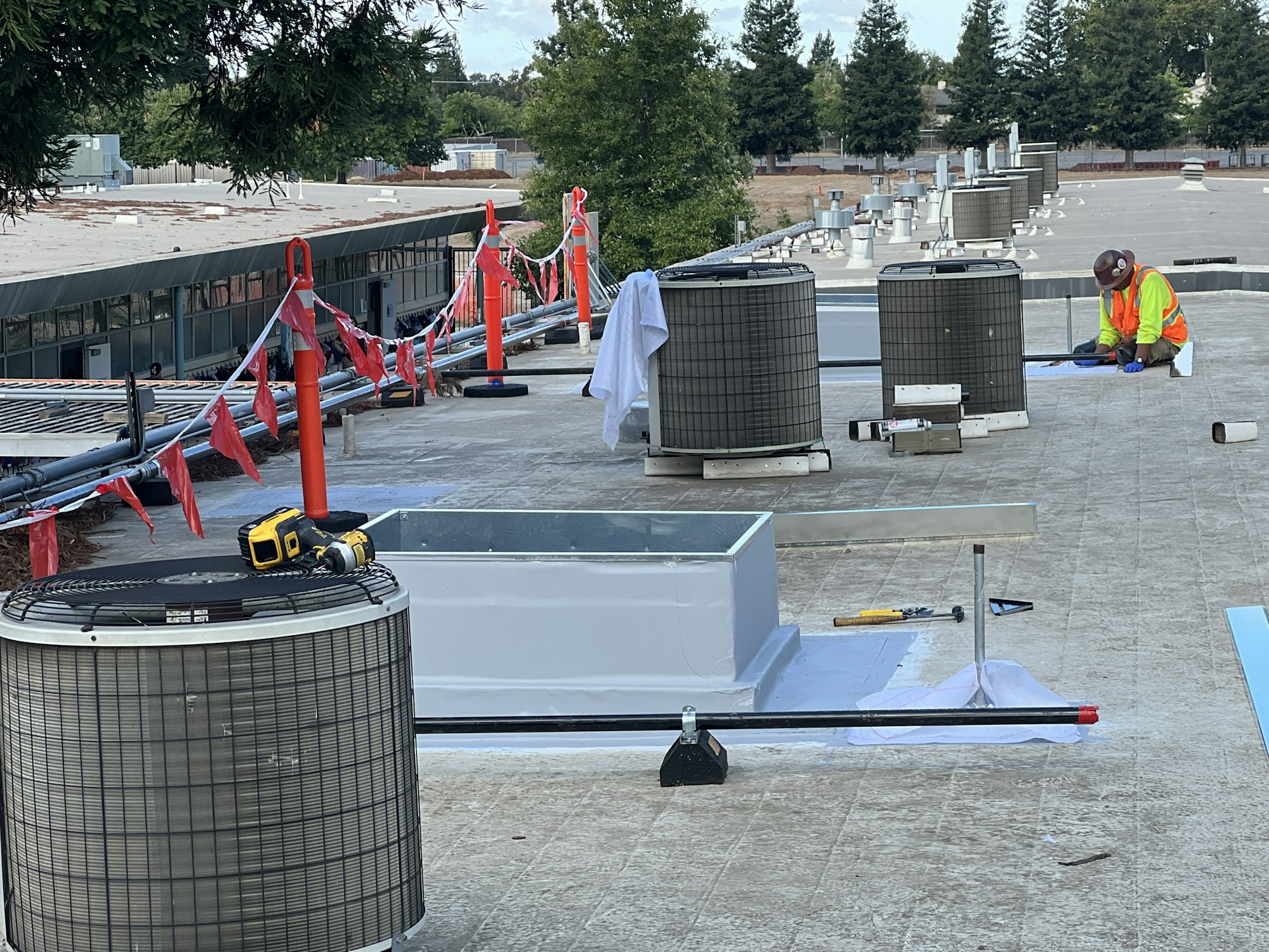 A worker in a safety vest is on a rooftop with multiple HVAC units, tools, and safety barriers.