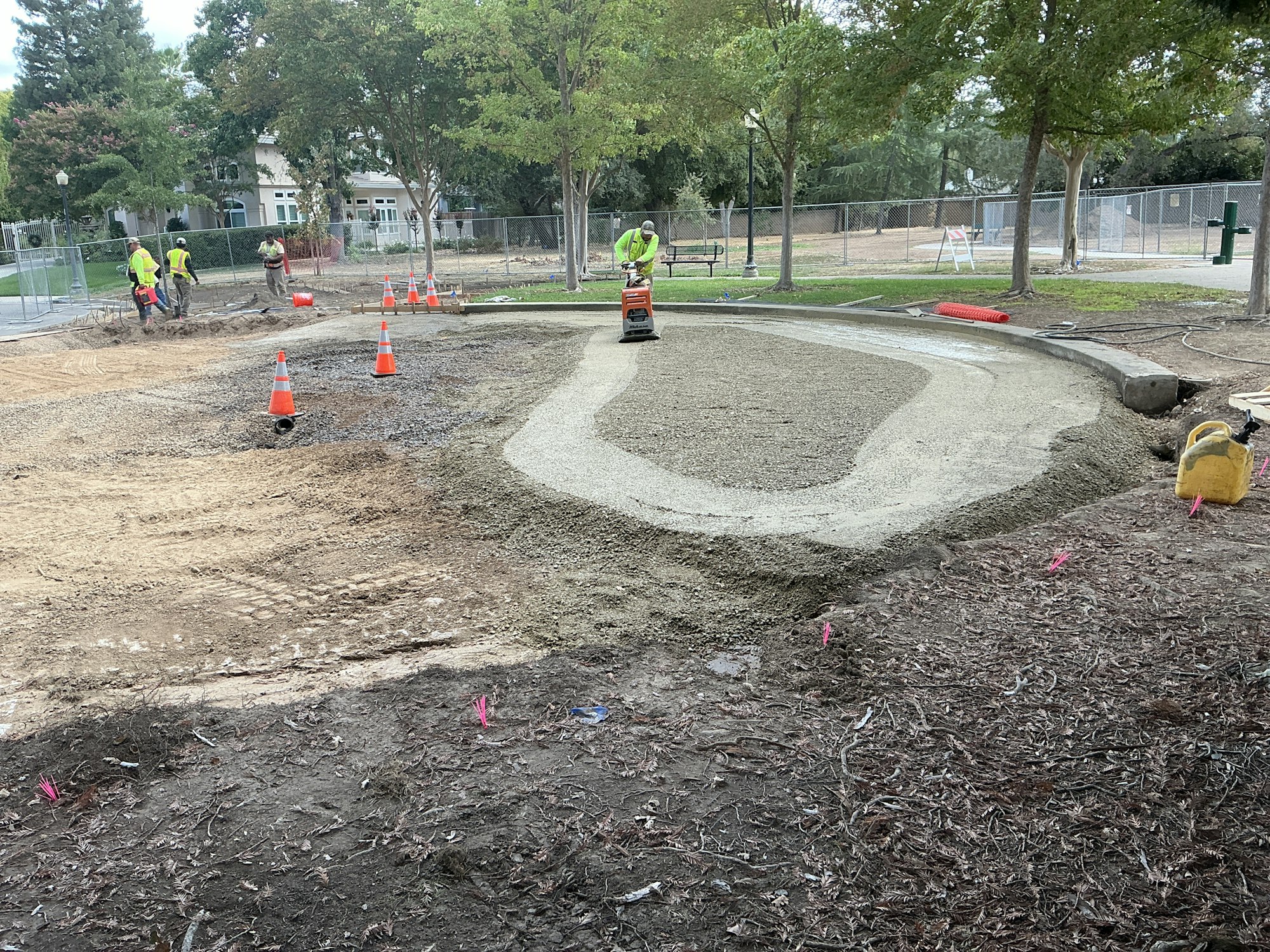 Construction site with workers preparing a foundation. Orange cones, dirt, and gravel are visible. Trees and a fence in the background.