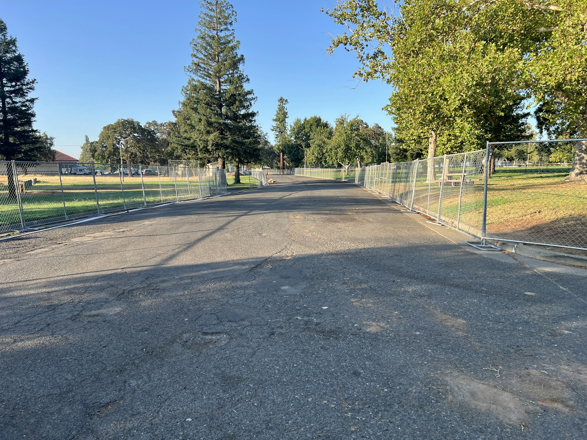 A paved road flanked by chain-link fences, with trees and grass on both sides under a clear blue sky.