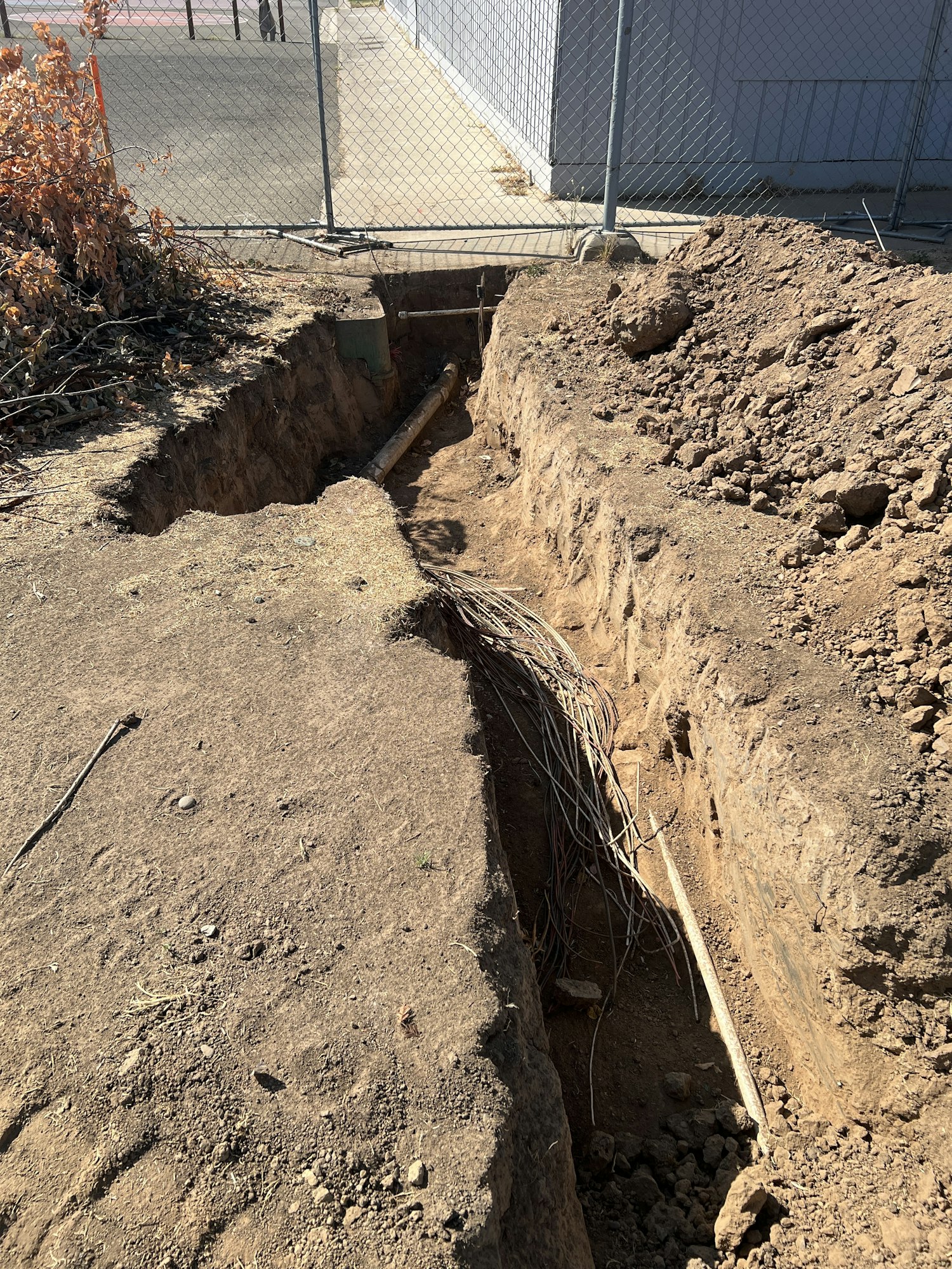 A dirt trench with exposed pipes and cables, surrounded by a fence and dried vegetation.