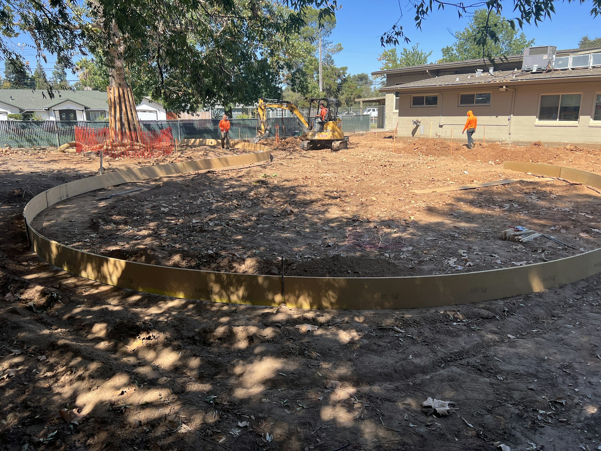 Construction site with circular formwork, workers in orange vests, earthmoving equipment, and surrounding trees and buildings.