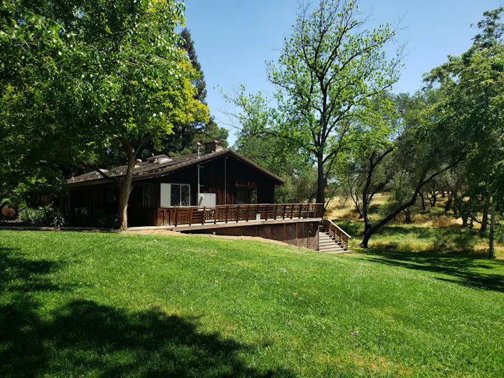 A rustic wooden house with a wooden deck, surrounded by green grass and trees under a clear blue sky.