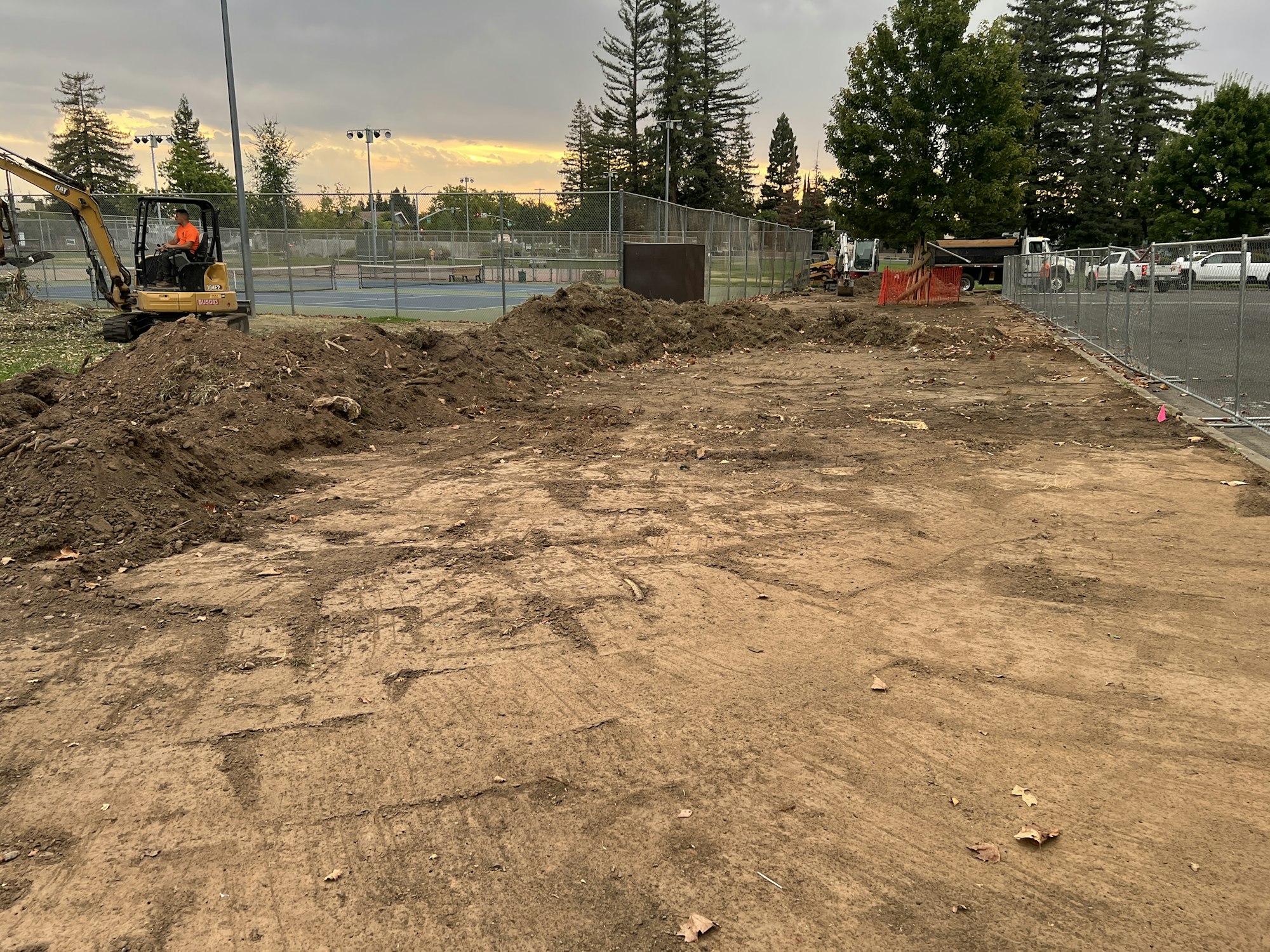 A construction site with dirt, a small excavator, and fencing is visible. In the background, there are tennis courts and trees.
