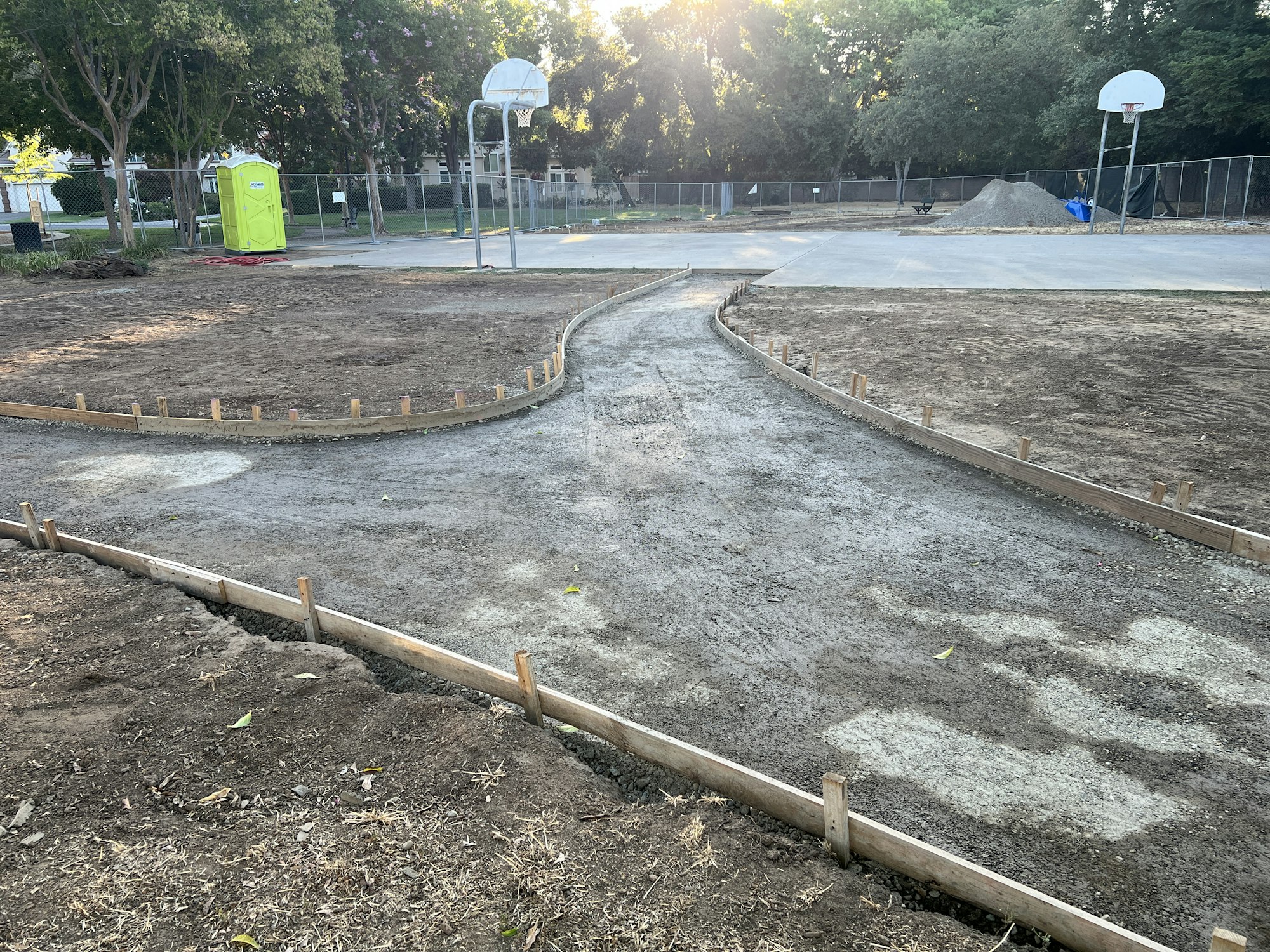 A construction site with a dirt path under construction, surrounded by wooden frames, near a basketball court and a portable toilet.