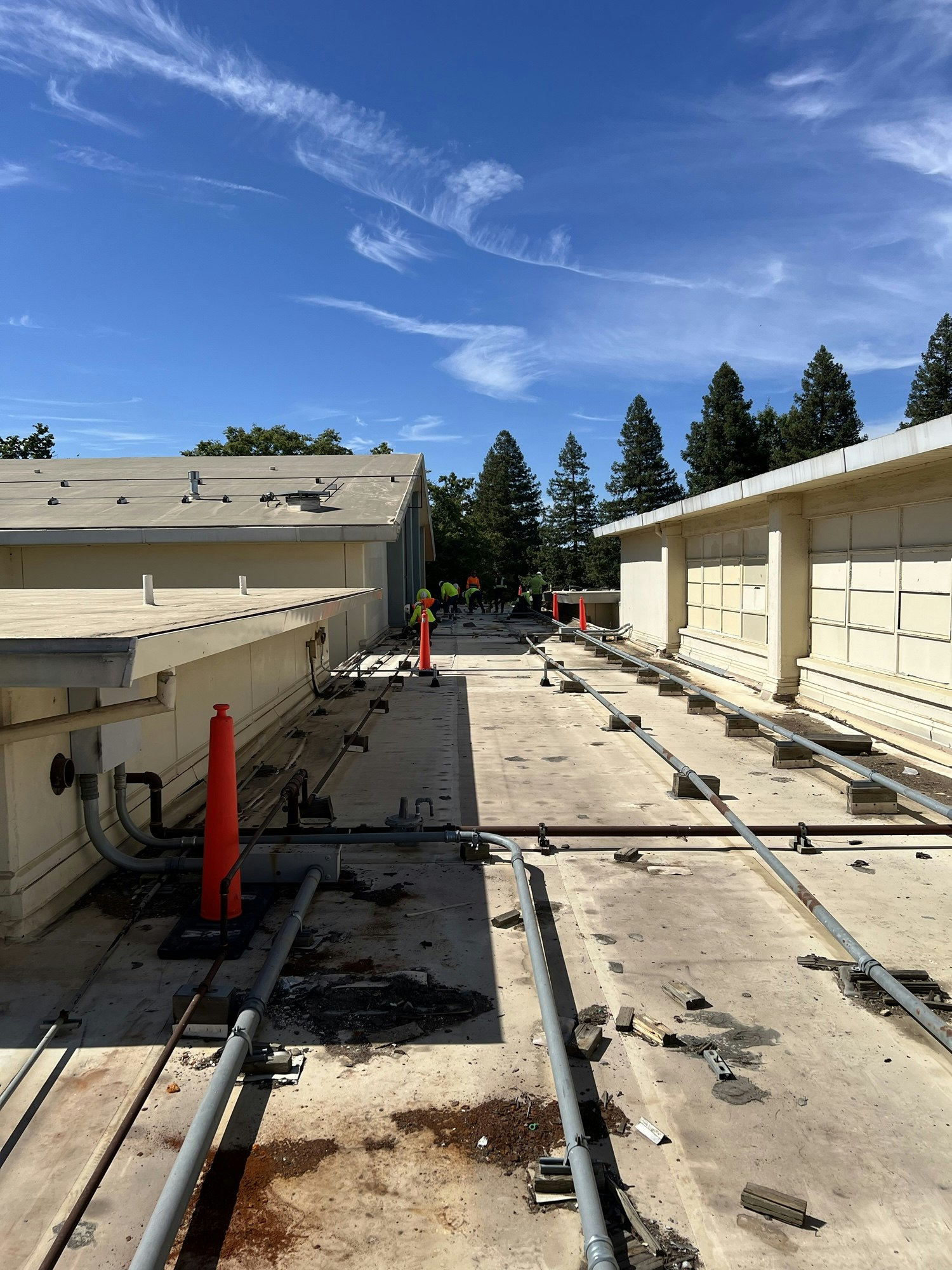 A rooftop with pipes, cones, and debris. Workers in the distance under a blue sky with clouds.