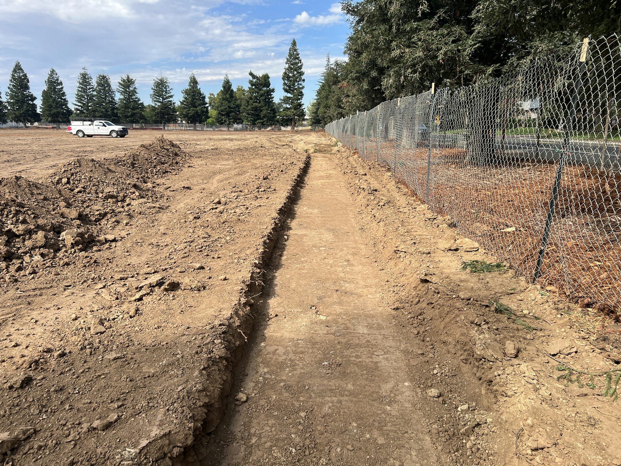 A dirt path next to a chain-link fence, with a white truck and trees in the background.
