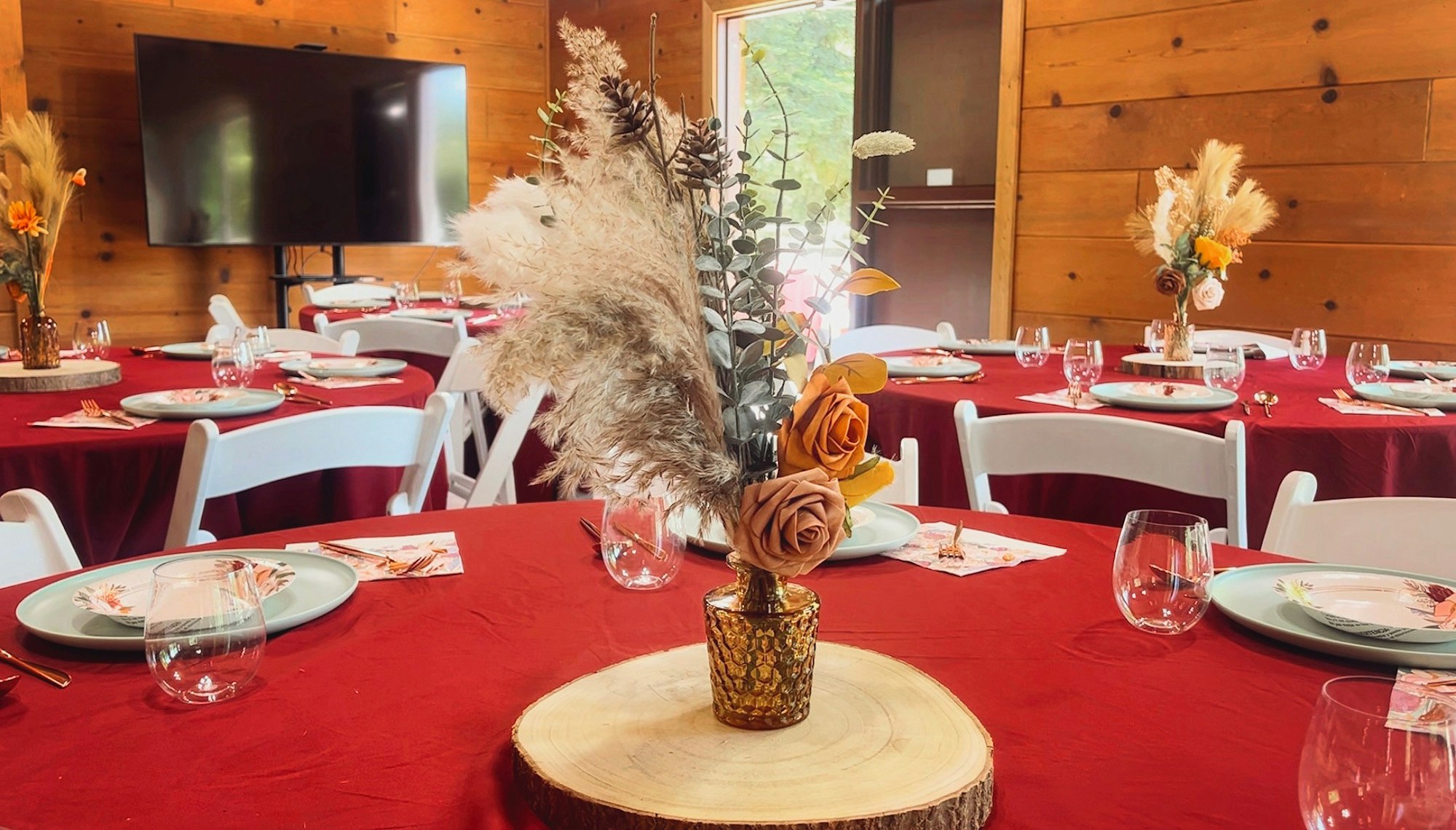 Elegant dining setup with red tablecloths, white chairs, rustic floral centerpieces, plates, and glasses arranged on wooden tables.