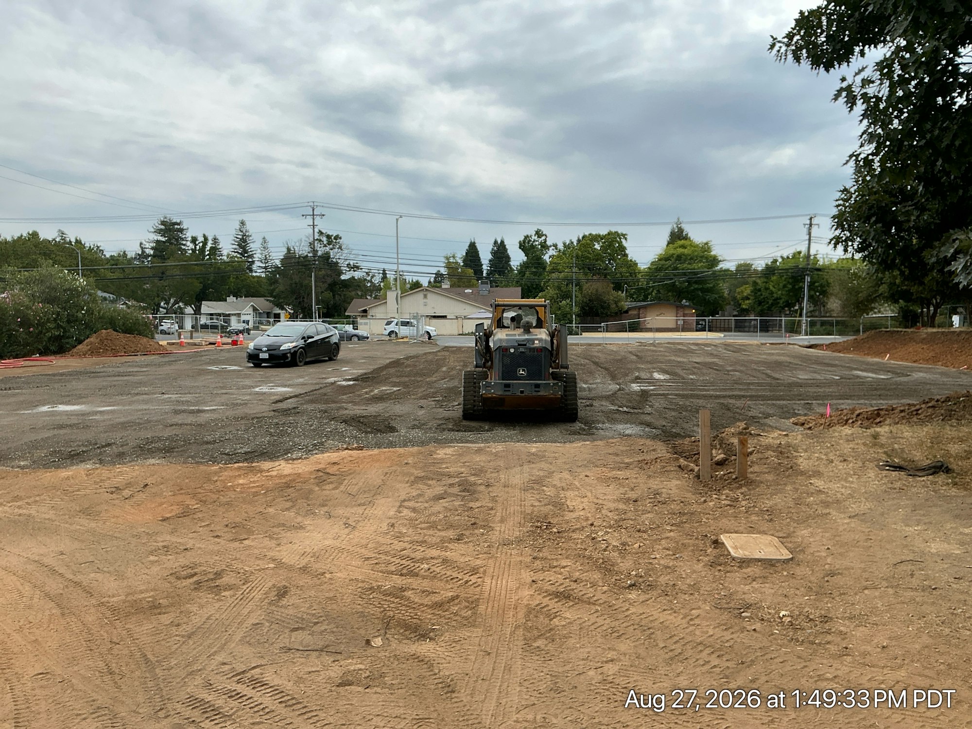 A construction site with heavy machinery, a parked car, and a cloudy sky in the background. Soil and gravel are visible.