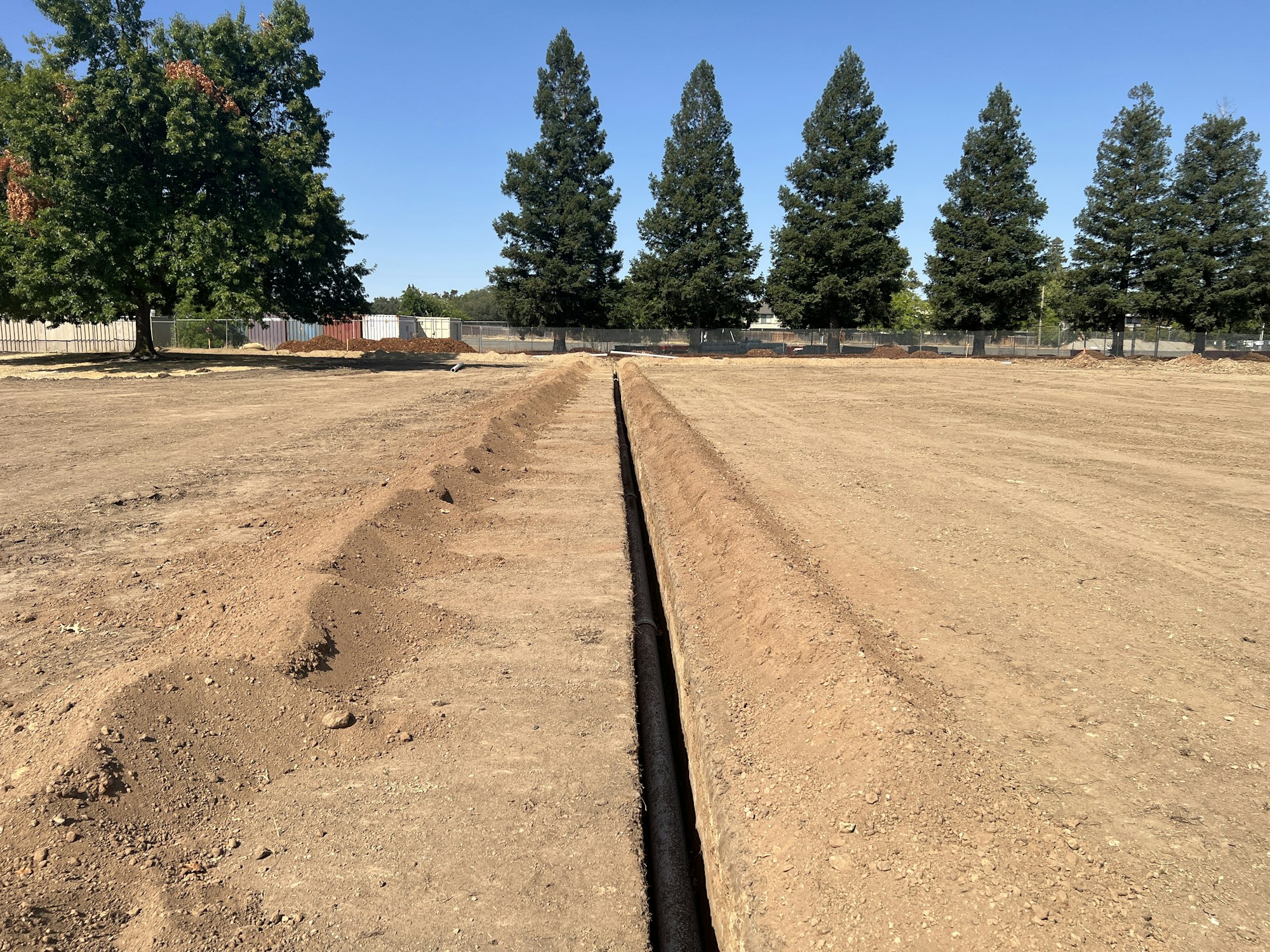 A long trench in a dirt field with trees in the background under a clear blue sky.