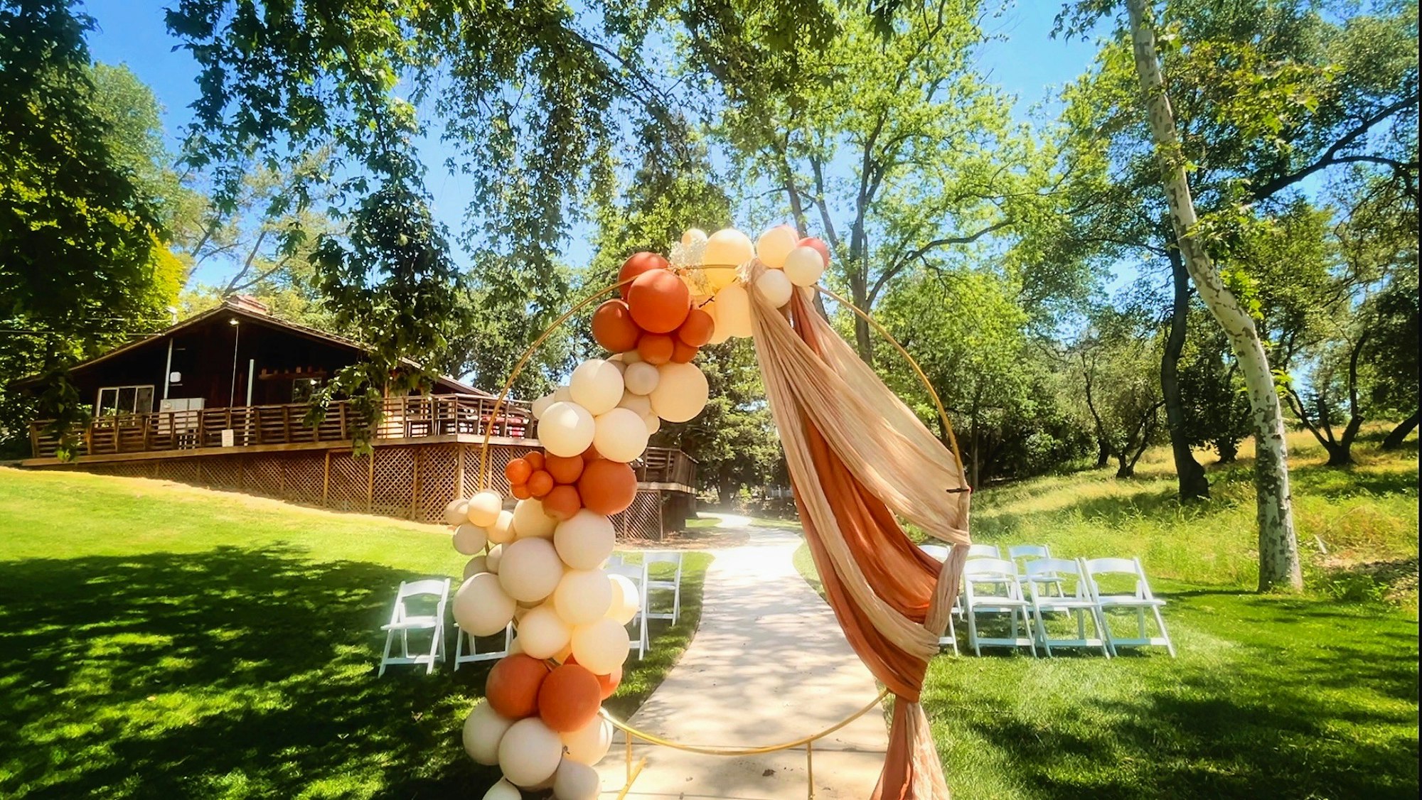 Wedding arch with balloons and drapery, outdoor setting with trees, grass, chairs, and a wooden building in the background.