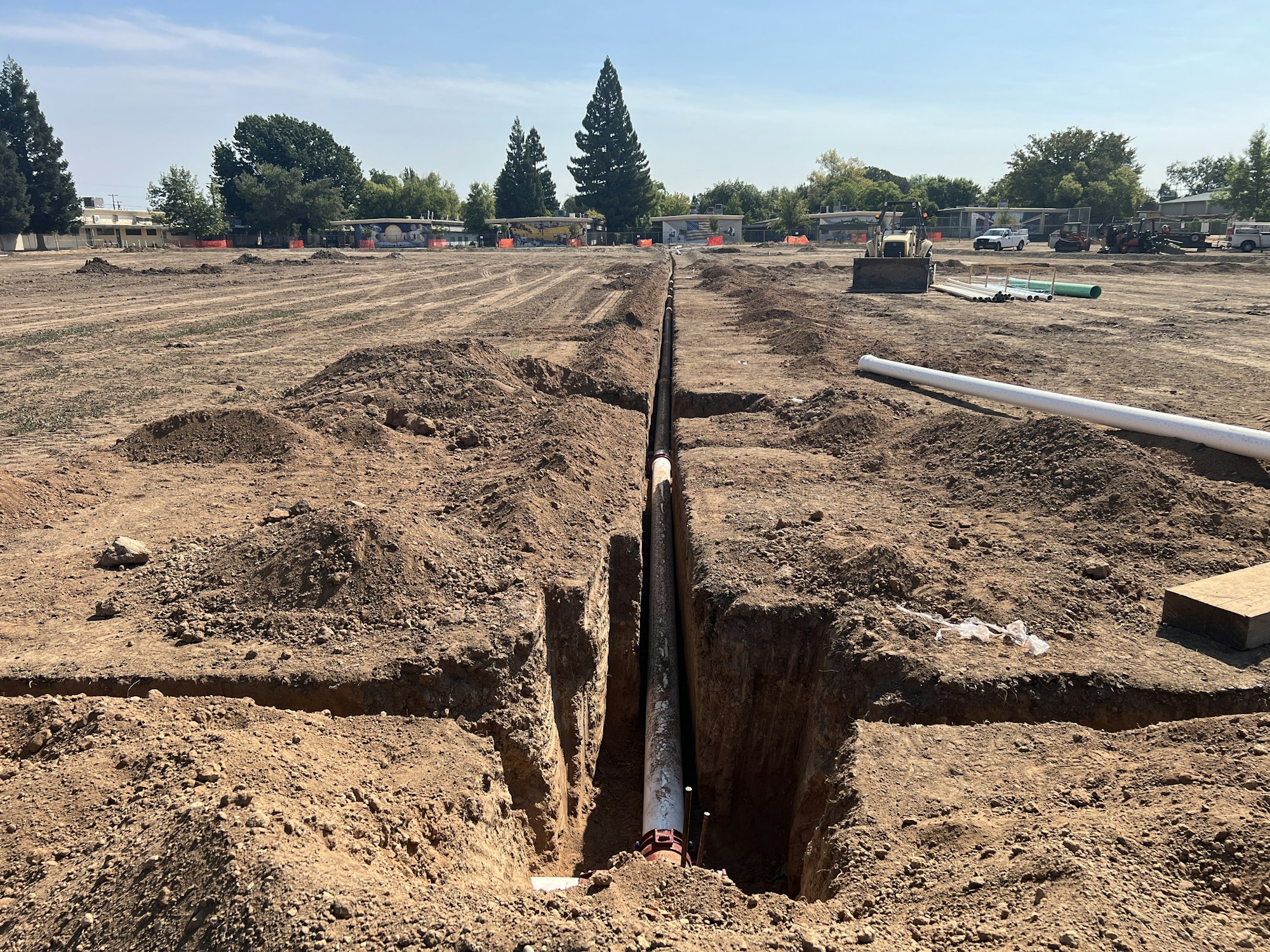 Construction site with a partially buried pipe in a trench, surrounded by dirt mounds and machinery in the background.