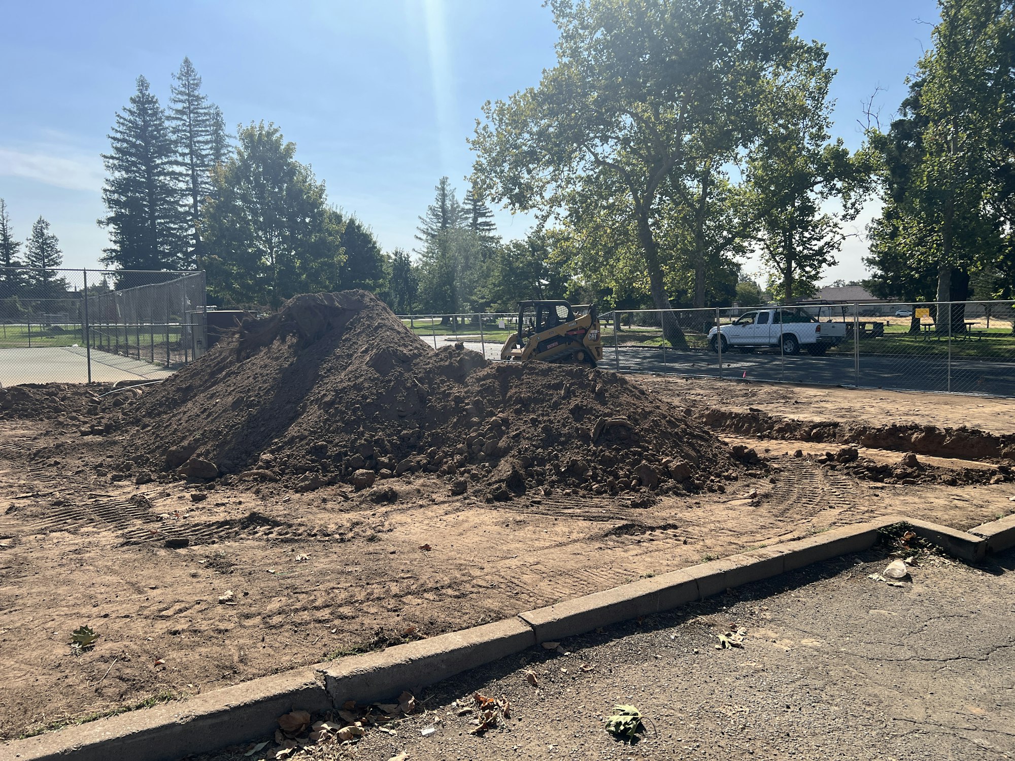 A construction site with a pile of dirt, a small bulldozer, and fencing. Trees and a vehicle in the background.
