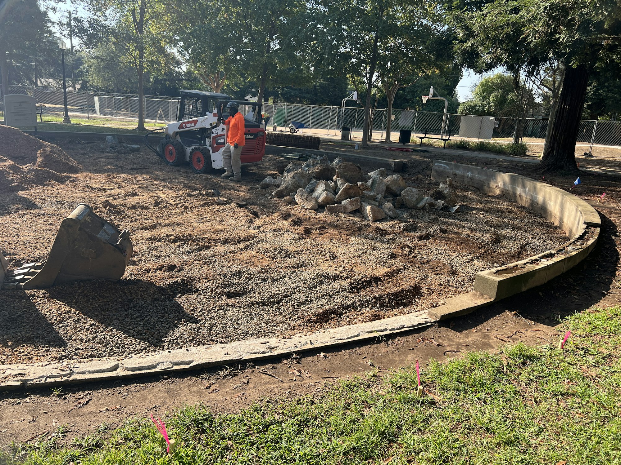 Construction site with a worker, pile of rocks, compact loader, and an excavator bucket on dirt and gravel area.