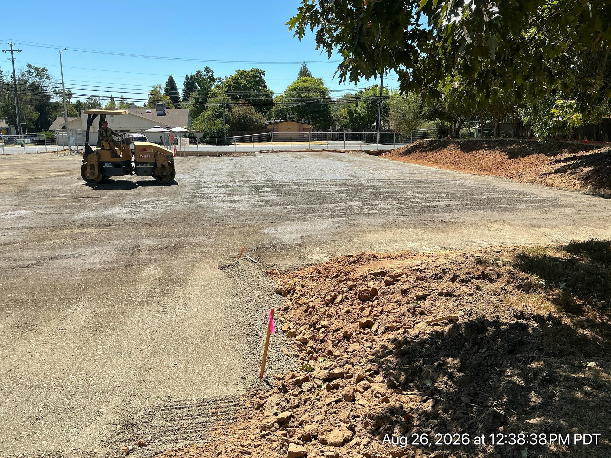 An empty construction site with a roller compacting gravel, surrounded by trees and marked boundaries.
