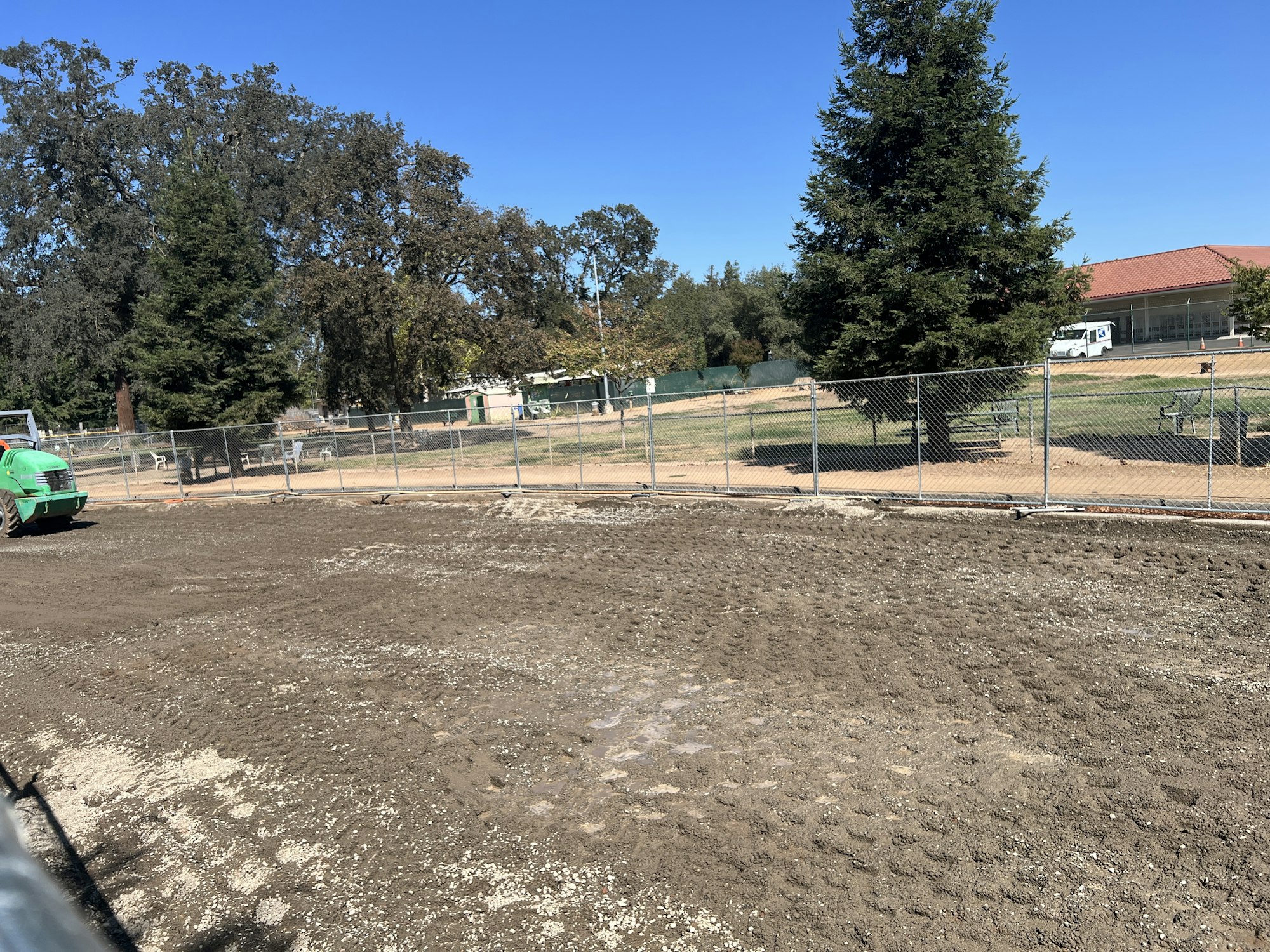 A fenced-off construction site with dirt, a green vehicle, trees, and buildings in the background.