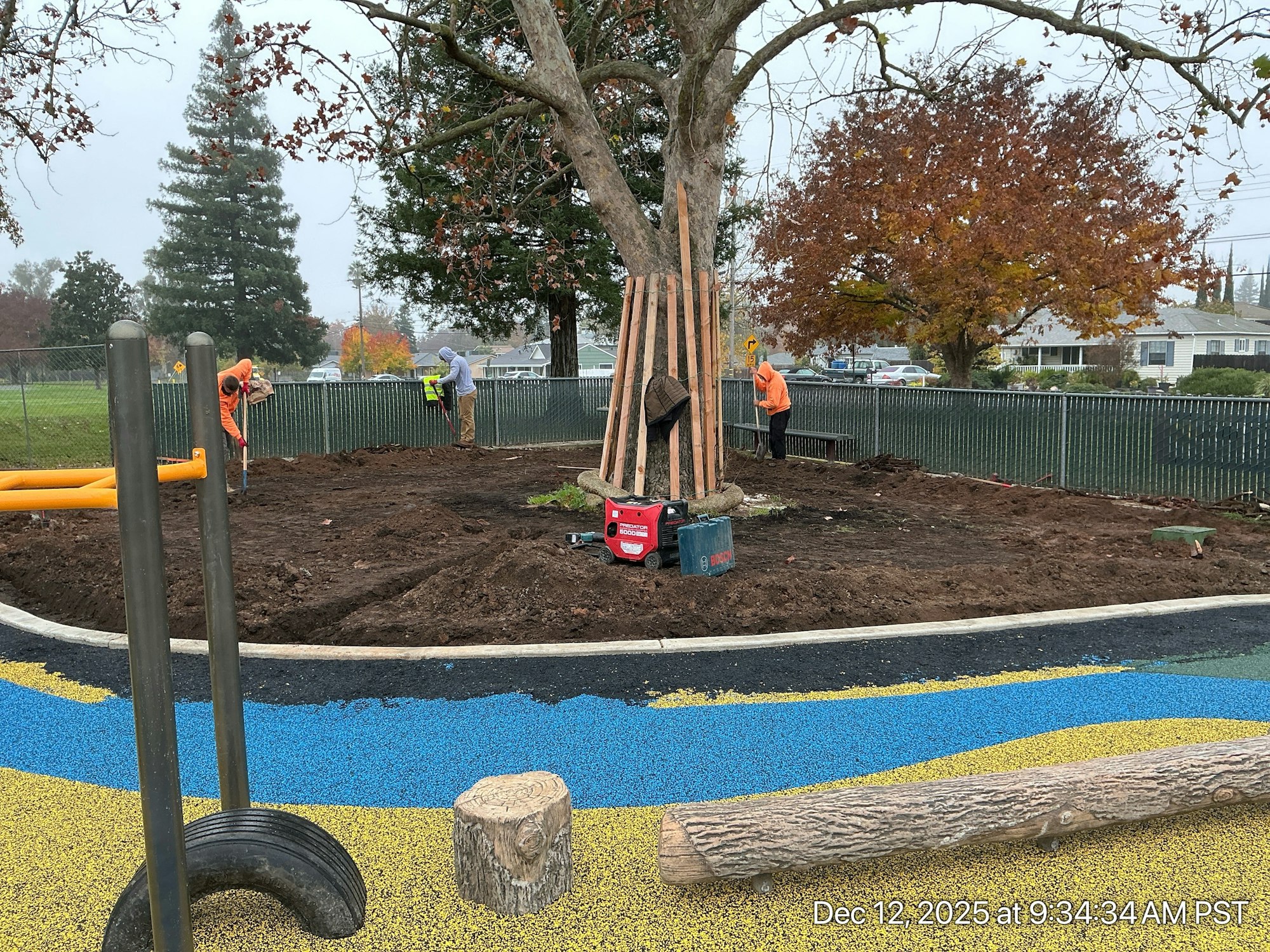 Workers are landscaping around a tree in a park, with colorful surfaces and tools visible in the area.