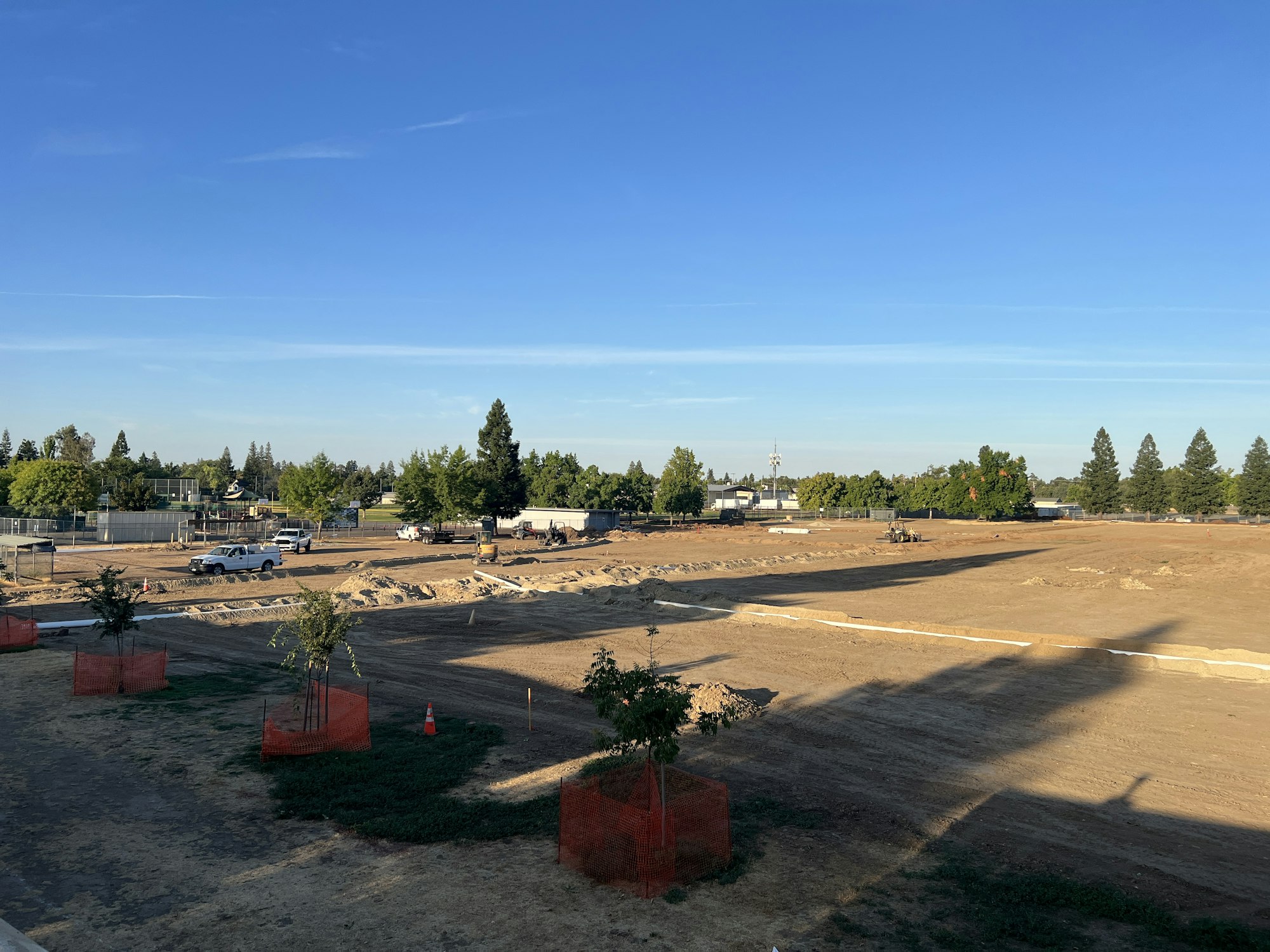 A construction site with vehicles, trees, and orange barriers on a clear day.
