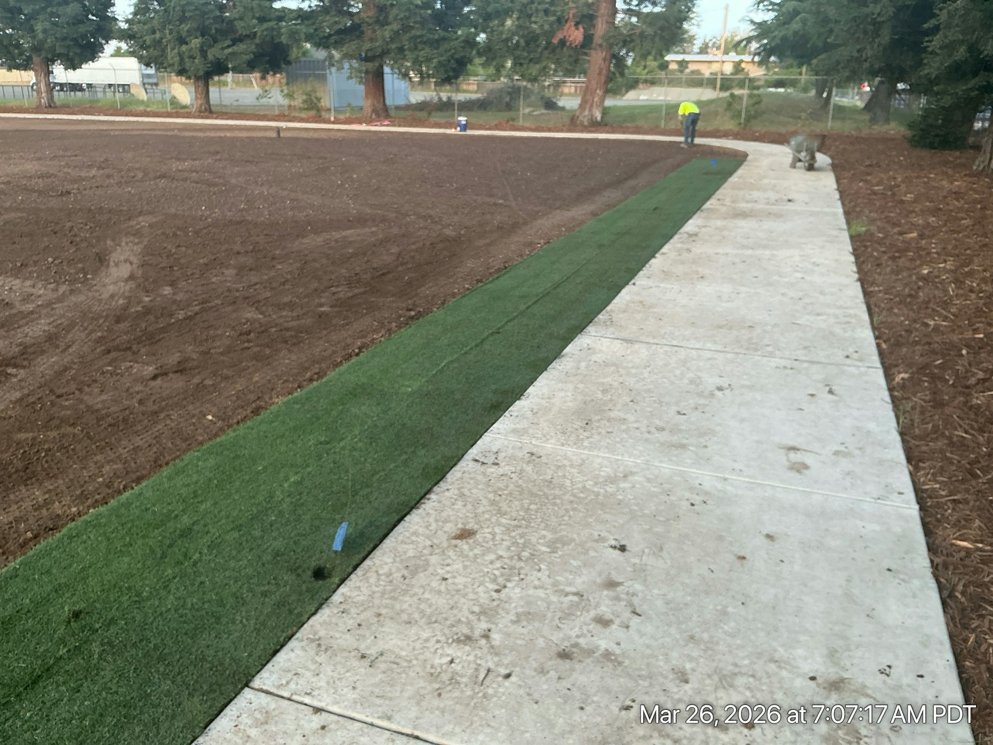 The image shows a construction site with freshly laid turf alongside a concrete path, under a tree-lined area.