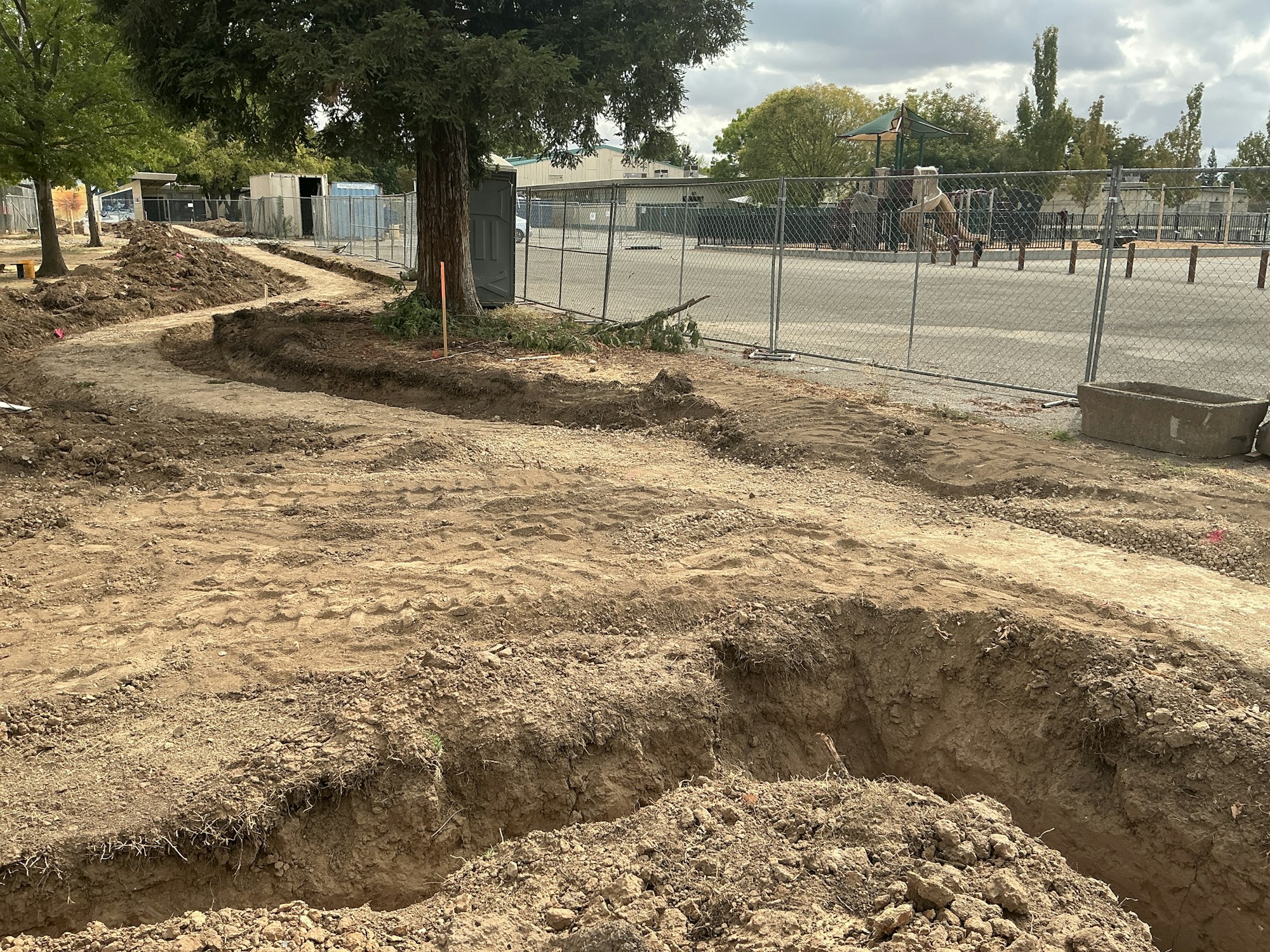 Construction site with dug trenches, fenced area, trees, and a playground in the background.