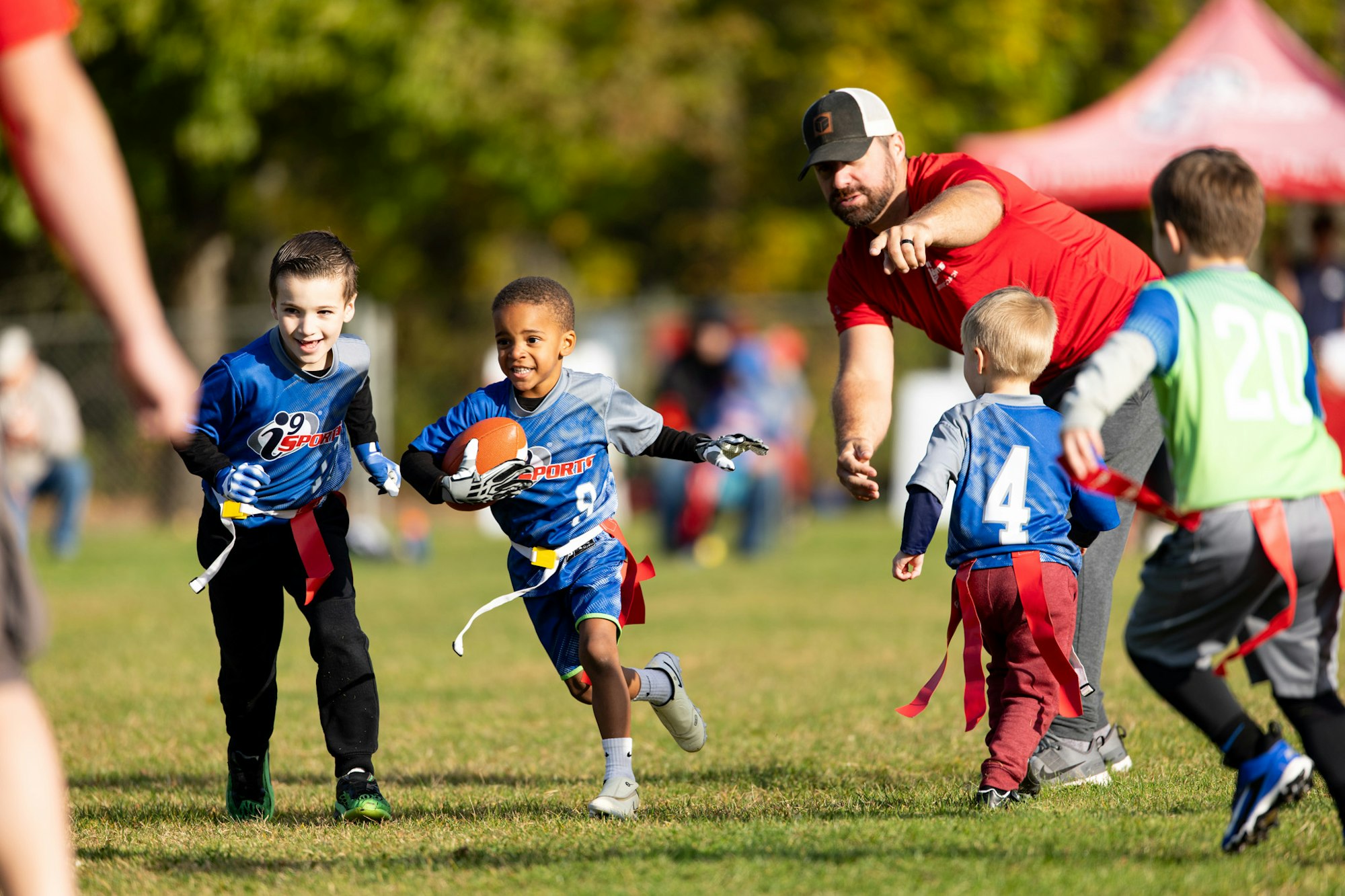 Kids playing flag football; a child runs with the ball, pursued by others. An adult in red gestures, watching the game.