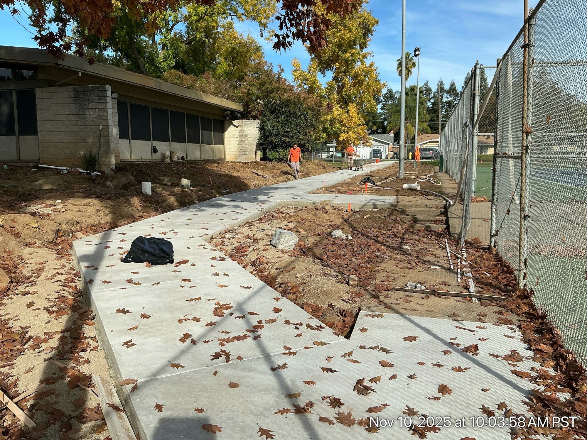 A construction site with a newly poured concrete sidewalk surrounded by fallen leaves and workers in bright orange shirts.