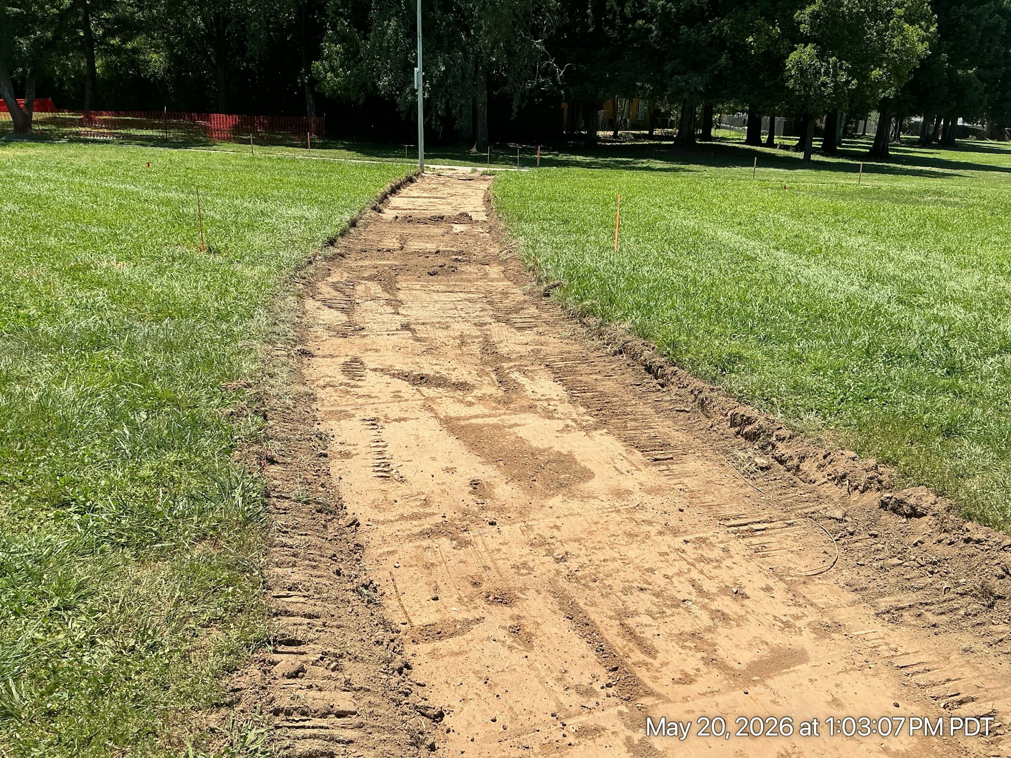 A dirt path is being constructed through a grassy area, with construction markers and a nearby light pole visible.
