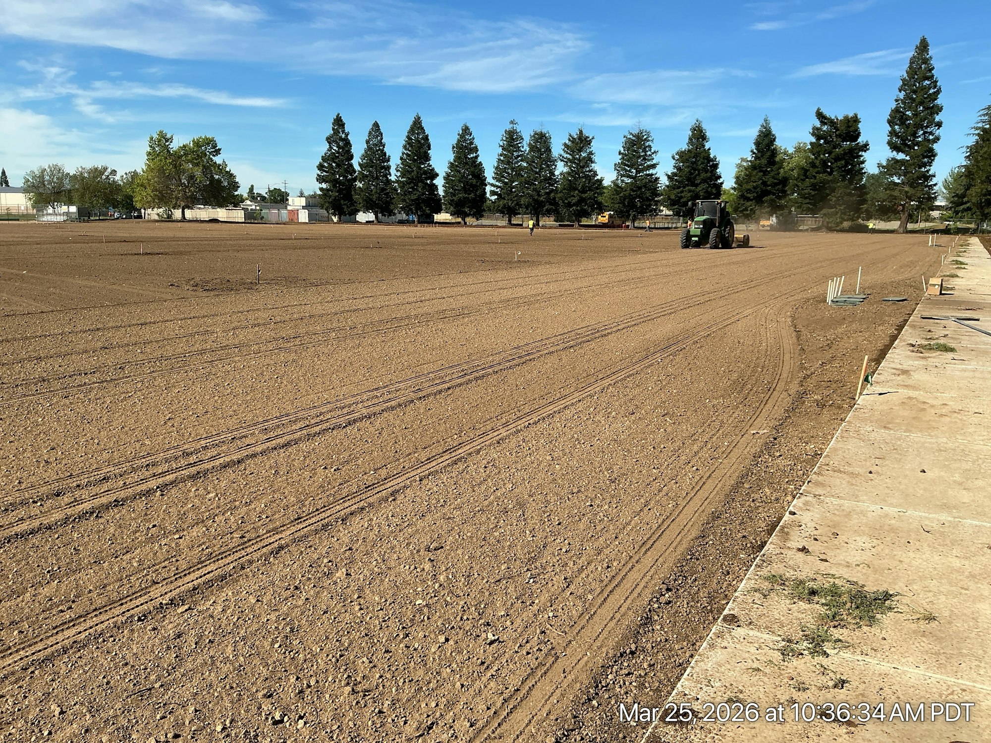 An agricultural field being worked on by a tractor, with clear skies and background trees visible.