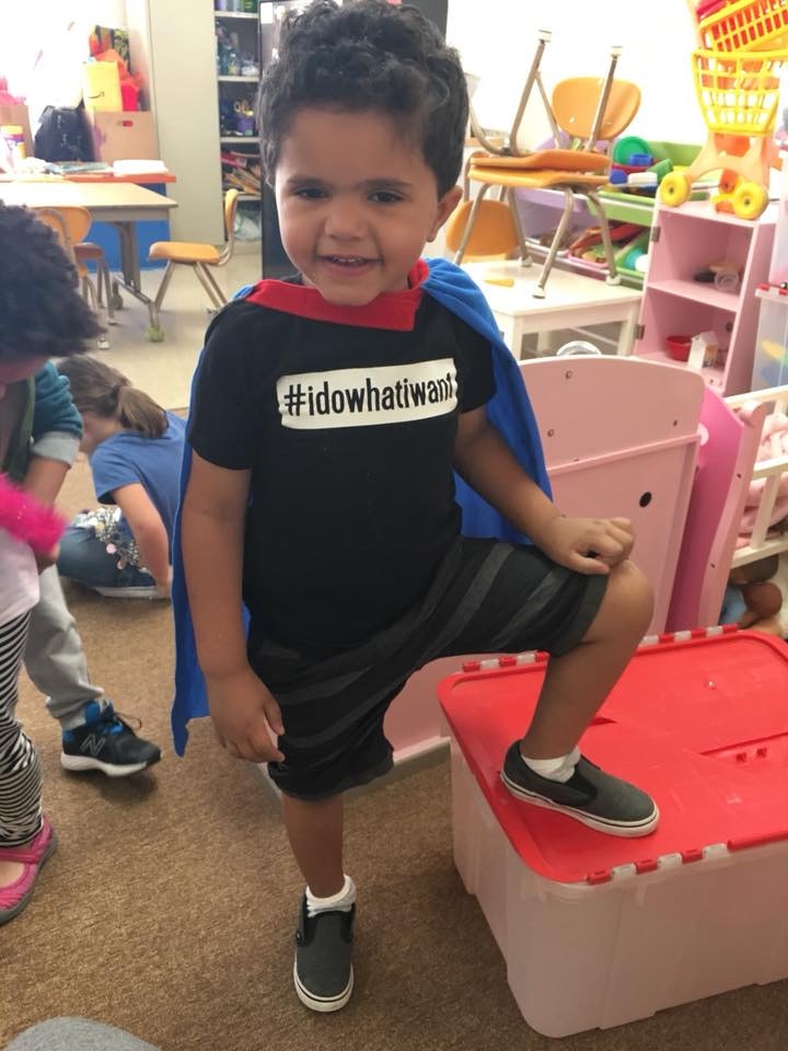 Tiny Tot child with a cape on and foot on a storage bin with toys all around him.