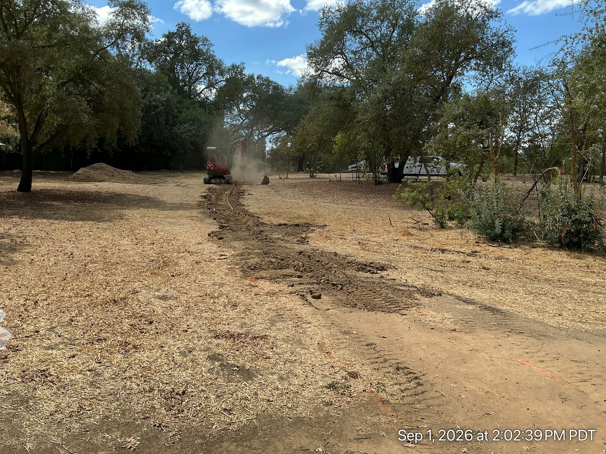 The image shows a dirt path being created with a machine in a dry area surrounded by trees, dust rising into the air.