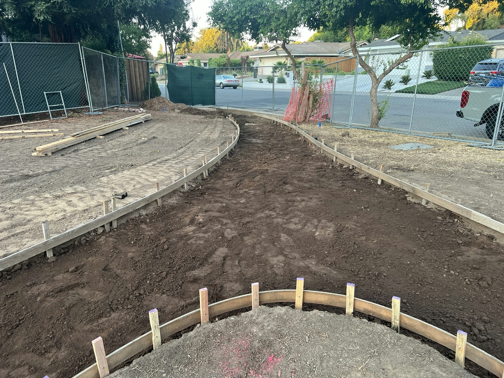 Construction site with curved wooden frames set in dirt, a fenced area, and trees in a residential neighborhood.