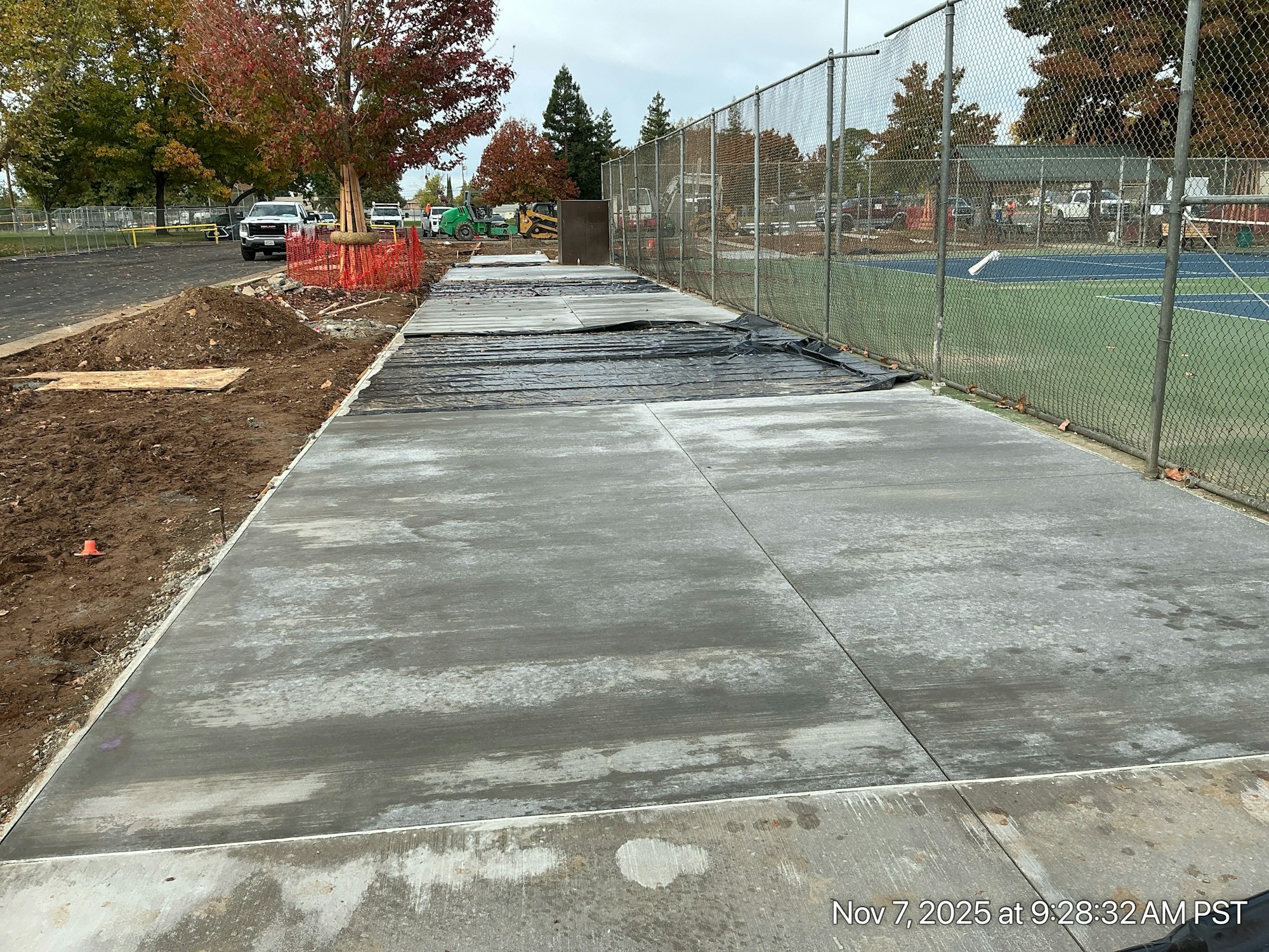 The image shows a newly poured concrete sidewalk under construction, surrounded by a fenced area with equipment and trees.