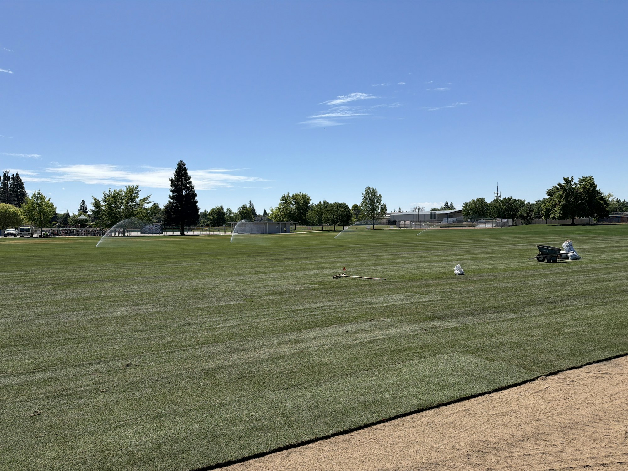 A sunny green sports field with sprinklers, trees, and maintenance equipment visible, under a clear blue sky.
