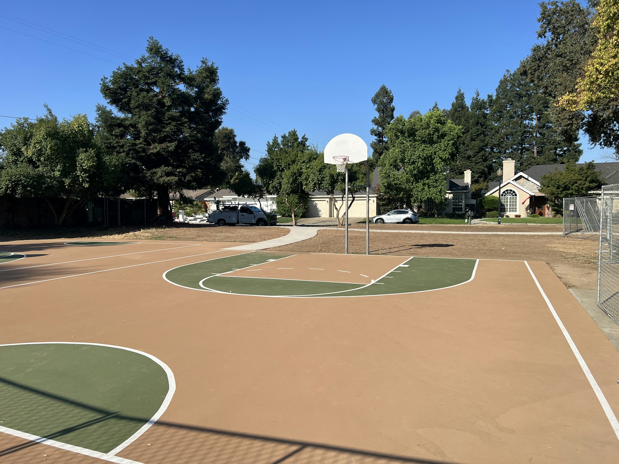 Outdoor basketball court with hoop, surrounded by trees and houses under a clear blue sky.