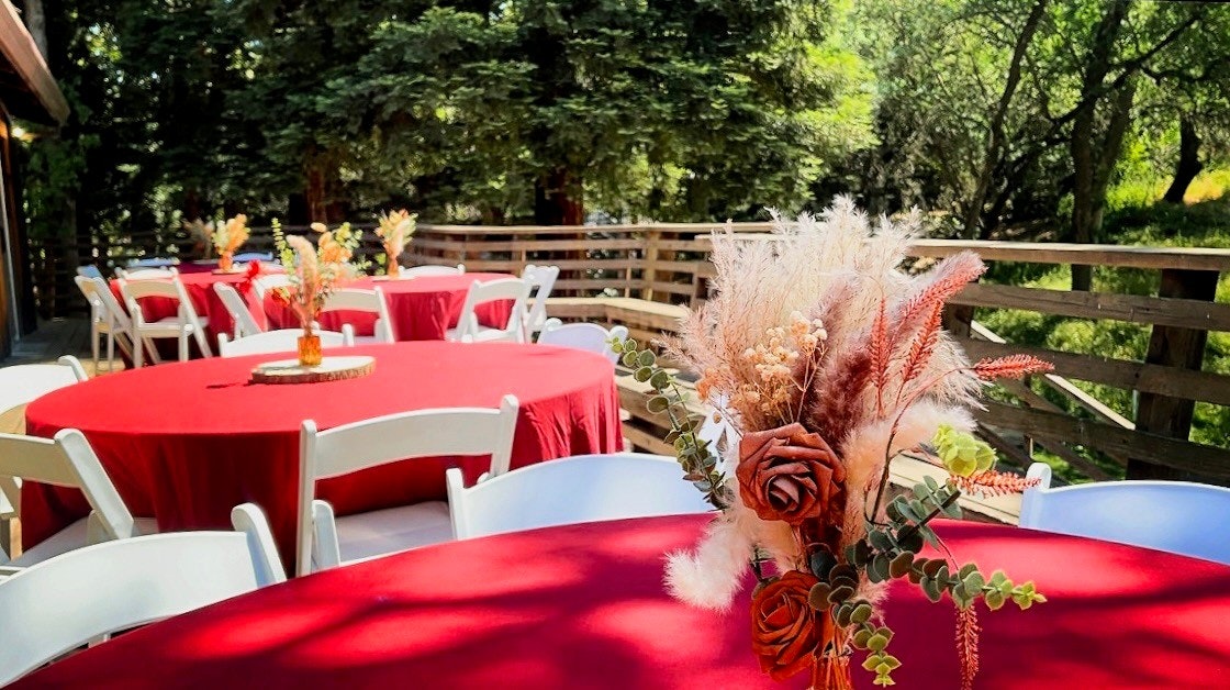Outdoor setting with tables covered in red cloths, white chairs, and floral centerpieces, surrounded by greenery.