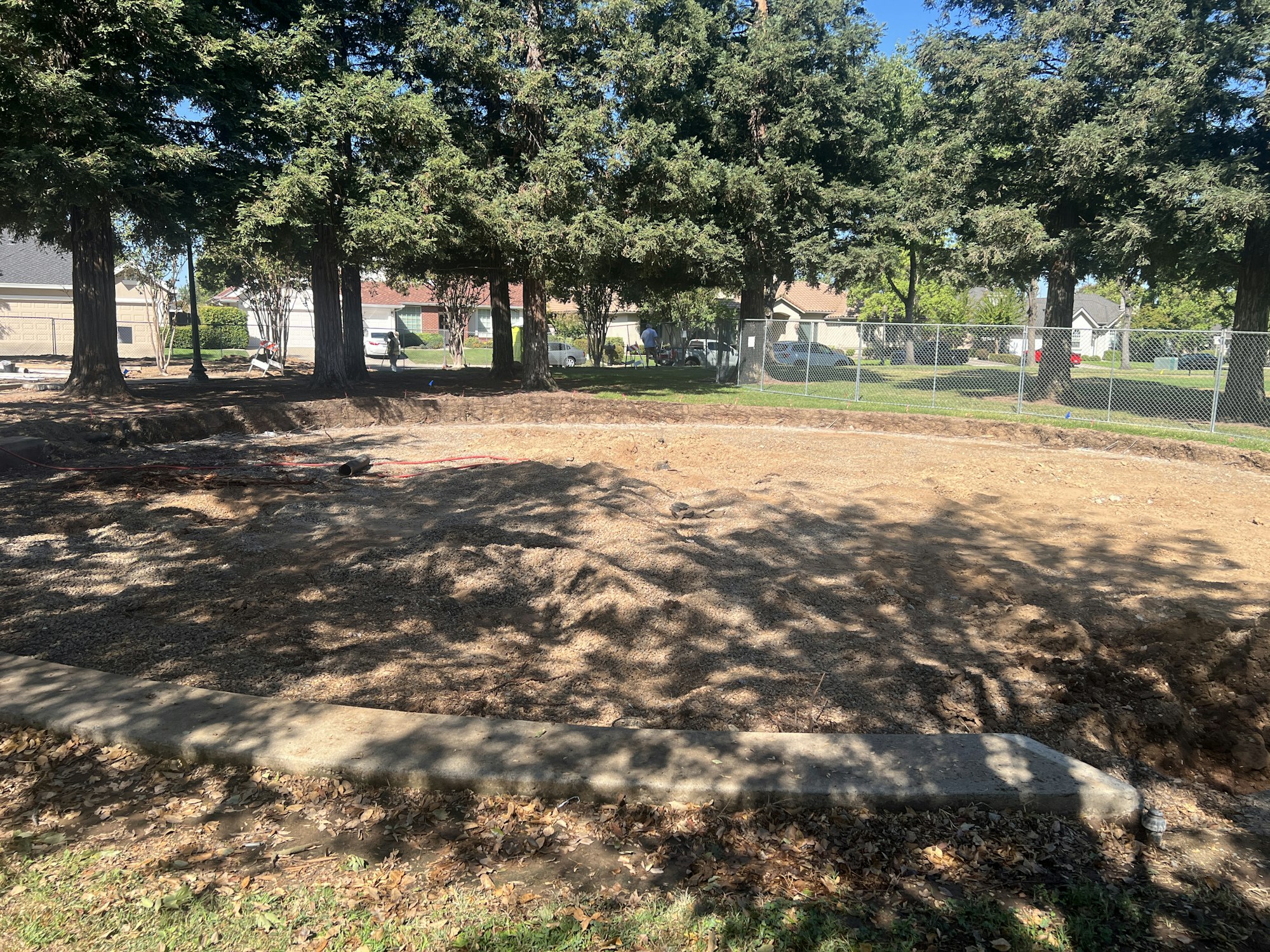 A construction site with cleared ground, surrounded by trees and a fence, near a residential area.