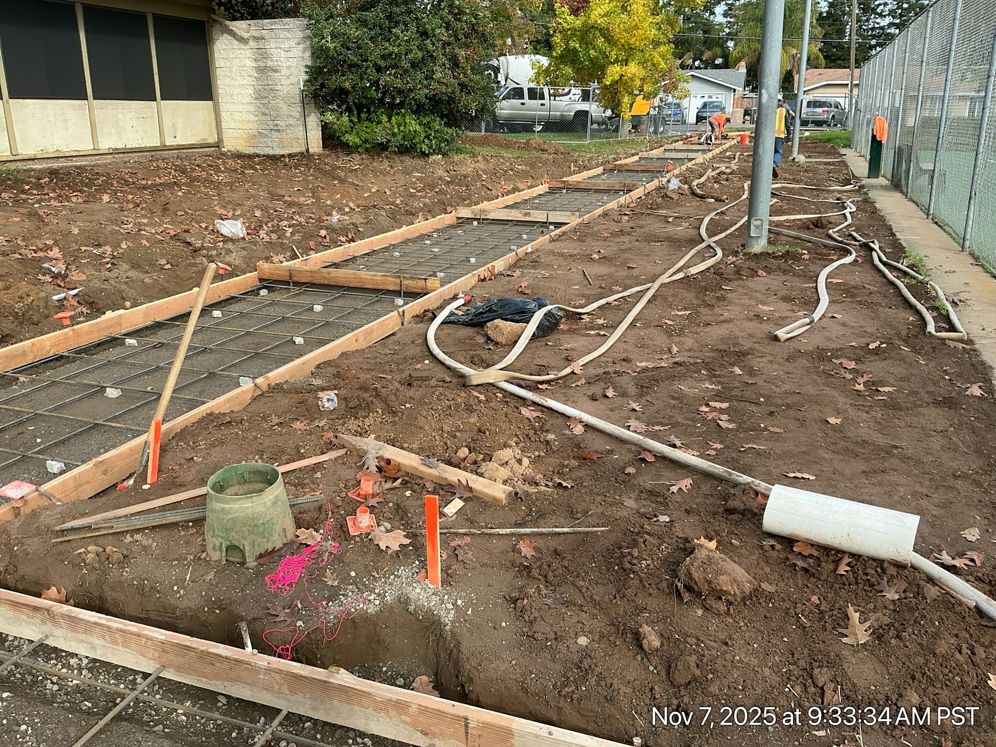 The image shows a construction site with wooden forms, rebar, dirt, and hoses, likely for laying concrete or landscaping.