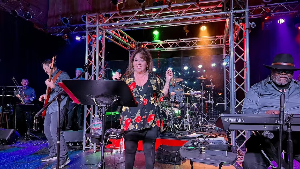 A lively music performance featuring a female vocalist, musicians on various instruments, and colorful stage lighting.