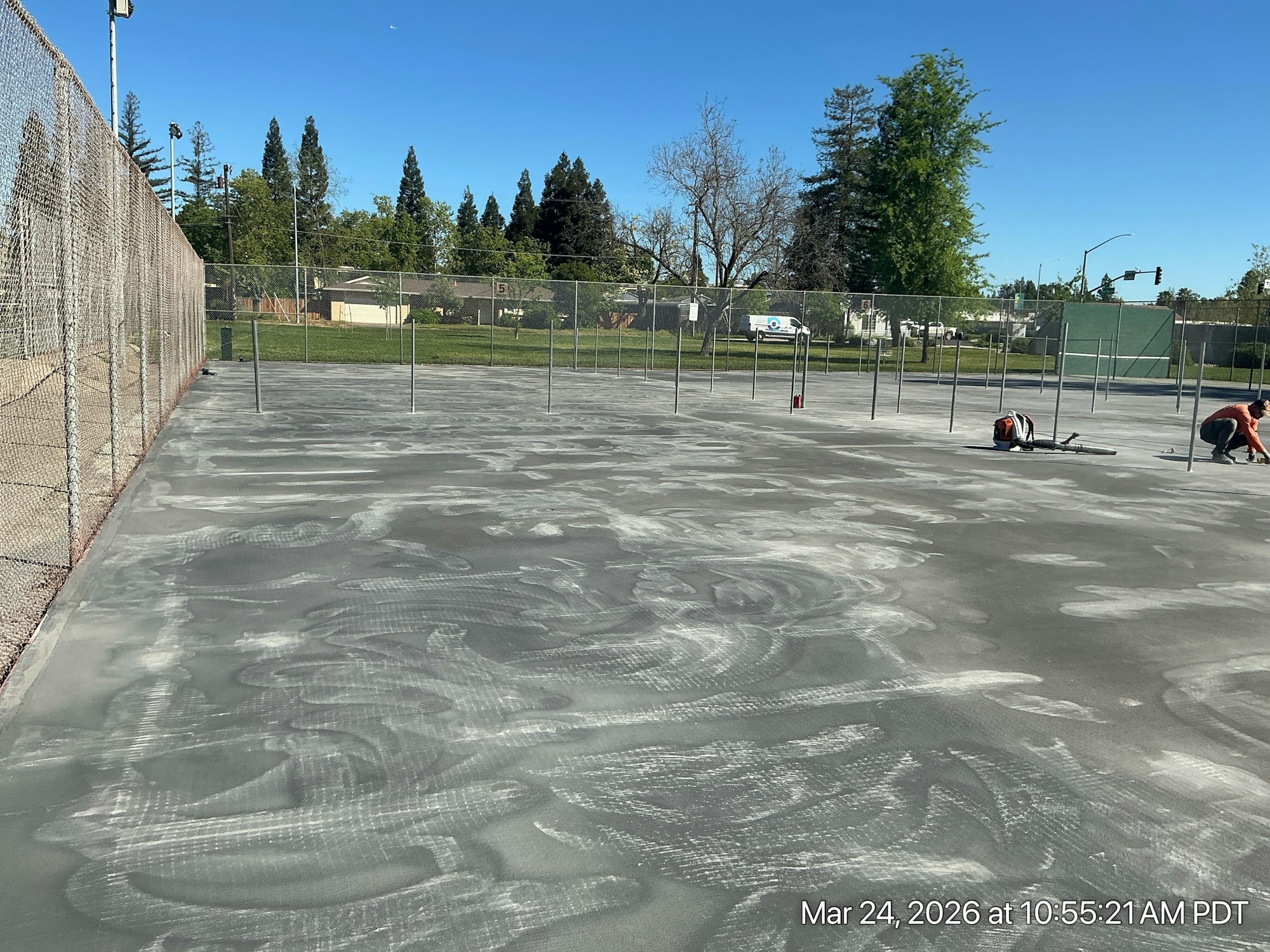 A gray court is being prepared or resurfaced, with workers and a fenced area nearby under a clear blue sky.