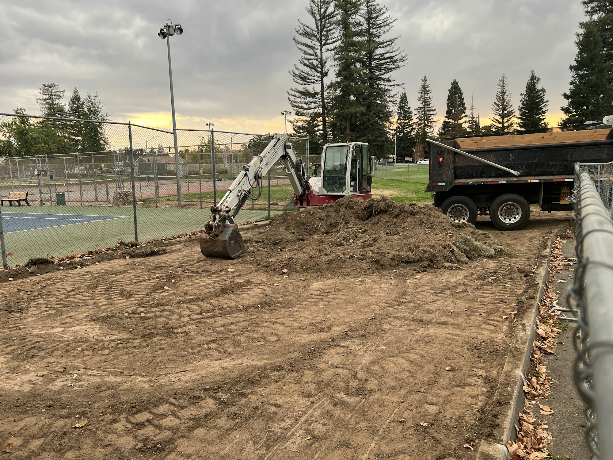 A small excavator and a dump truck near a pile of dirt beside tennis courts, with trees and a cloudy sky in the background.