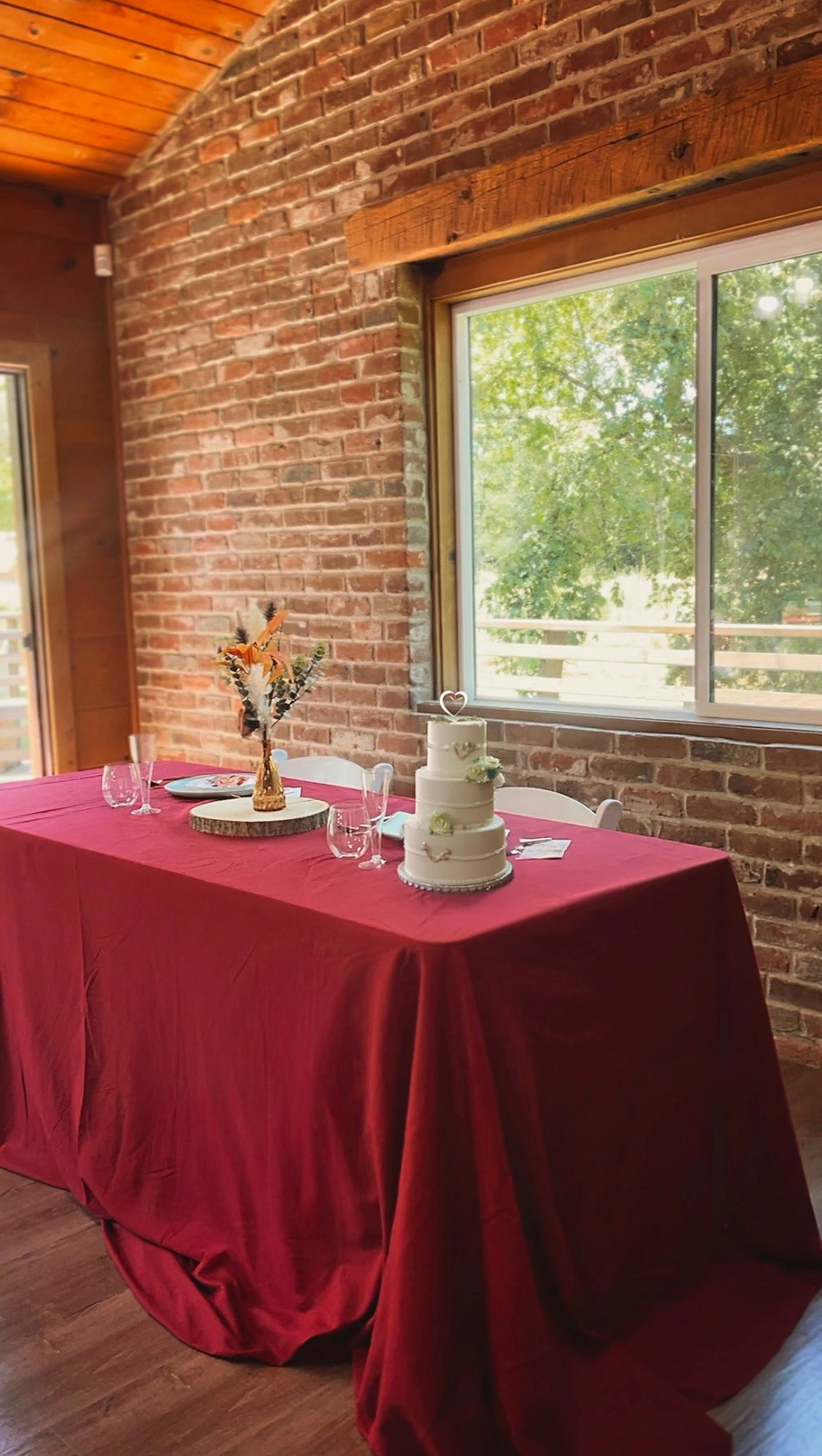A red tablecloth-covered table with a floral arrangement, a white cake, and glassware, set near a brick wall and large window.