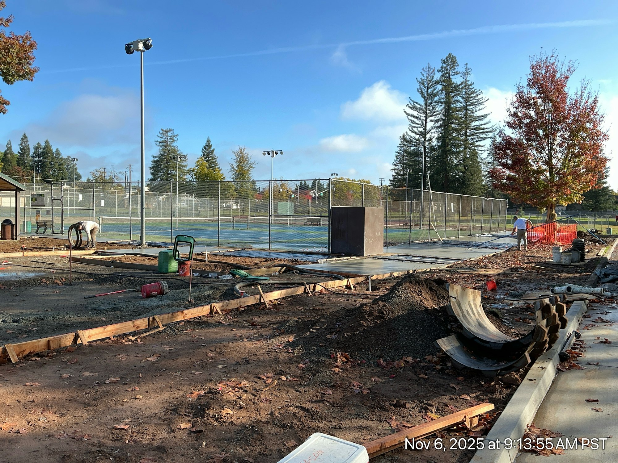 A construction site near tennis courts, with workers, tools, and materials visible under a clear sky.