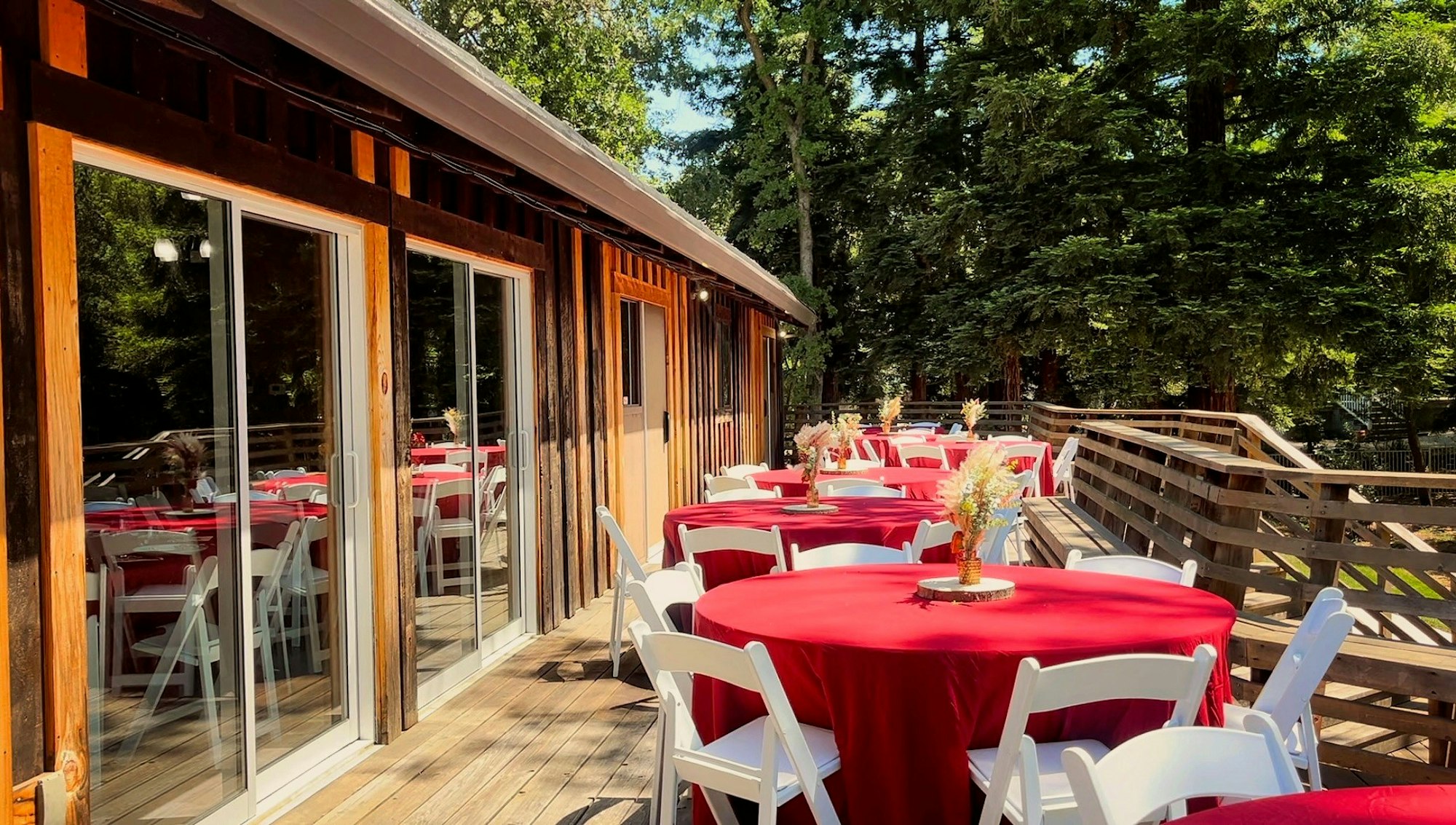 Outdoor deck with red tablecloths on round tables, white chairs, and floral centerpieces, next to a wooden building and trees.