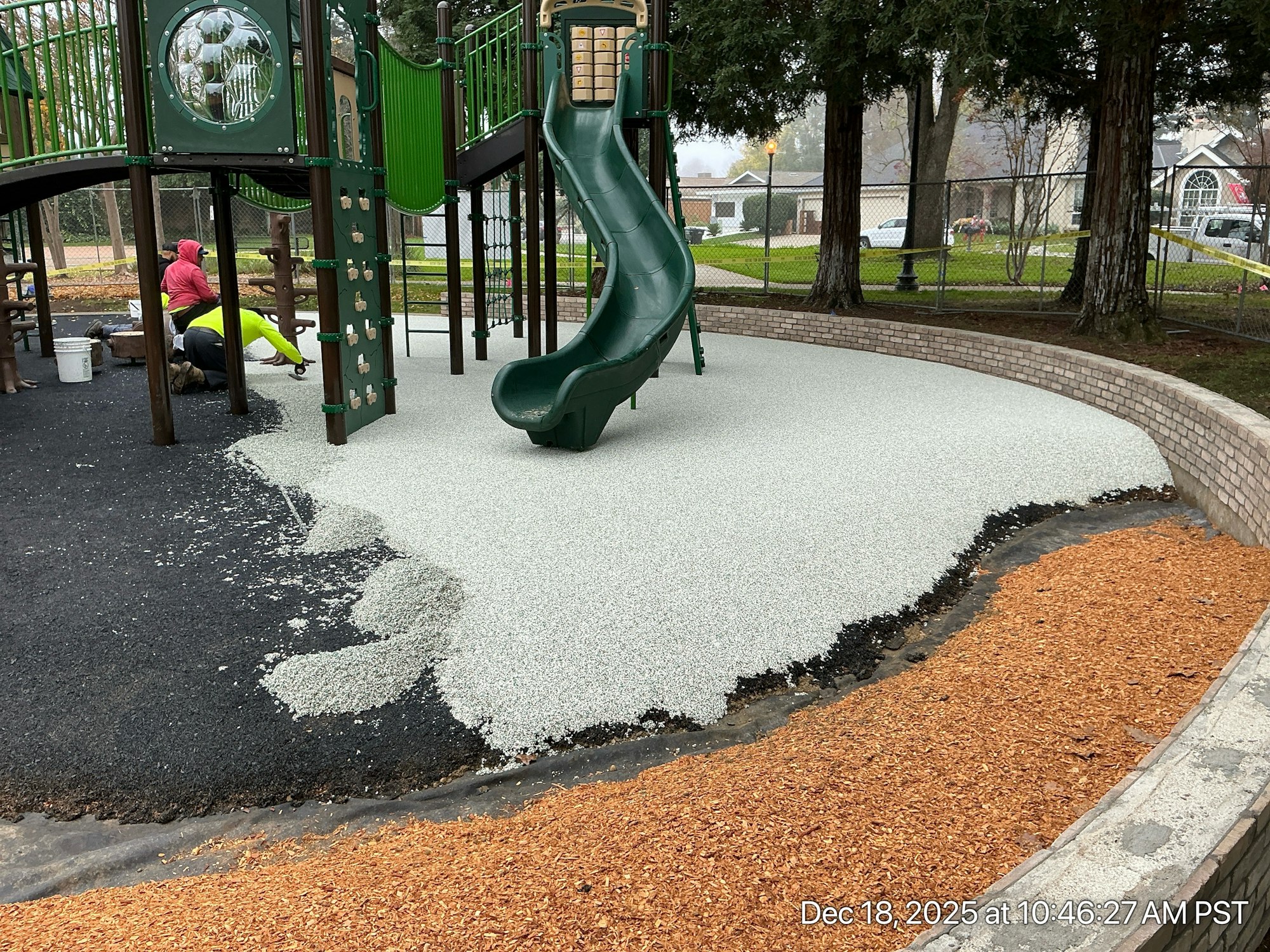The image shows a playground under construction, with new rubber surfacing being installed around a slide.