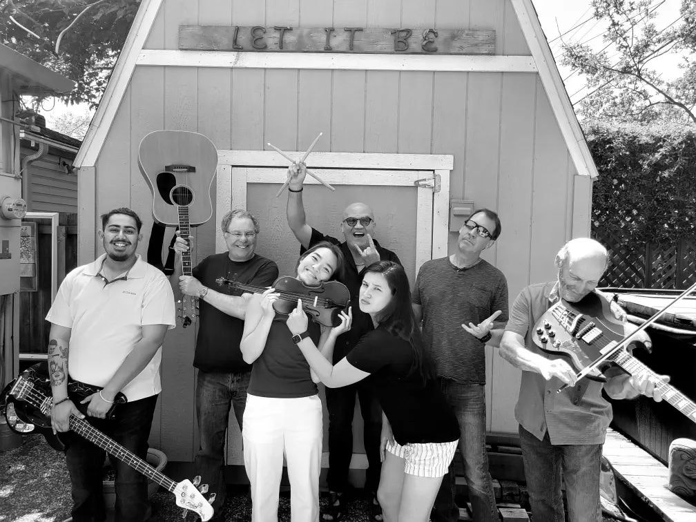 Seven people posing playfully with musical instruments in front of a shed with a "Let It Be" sign.