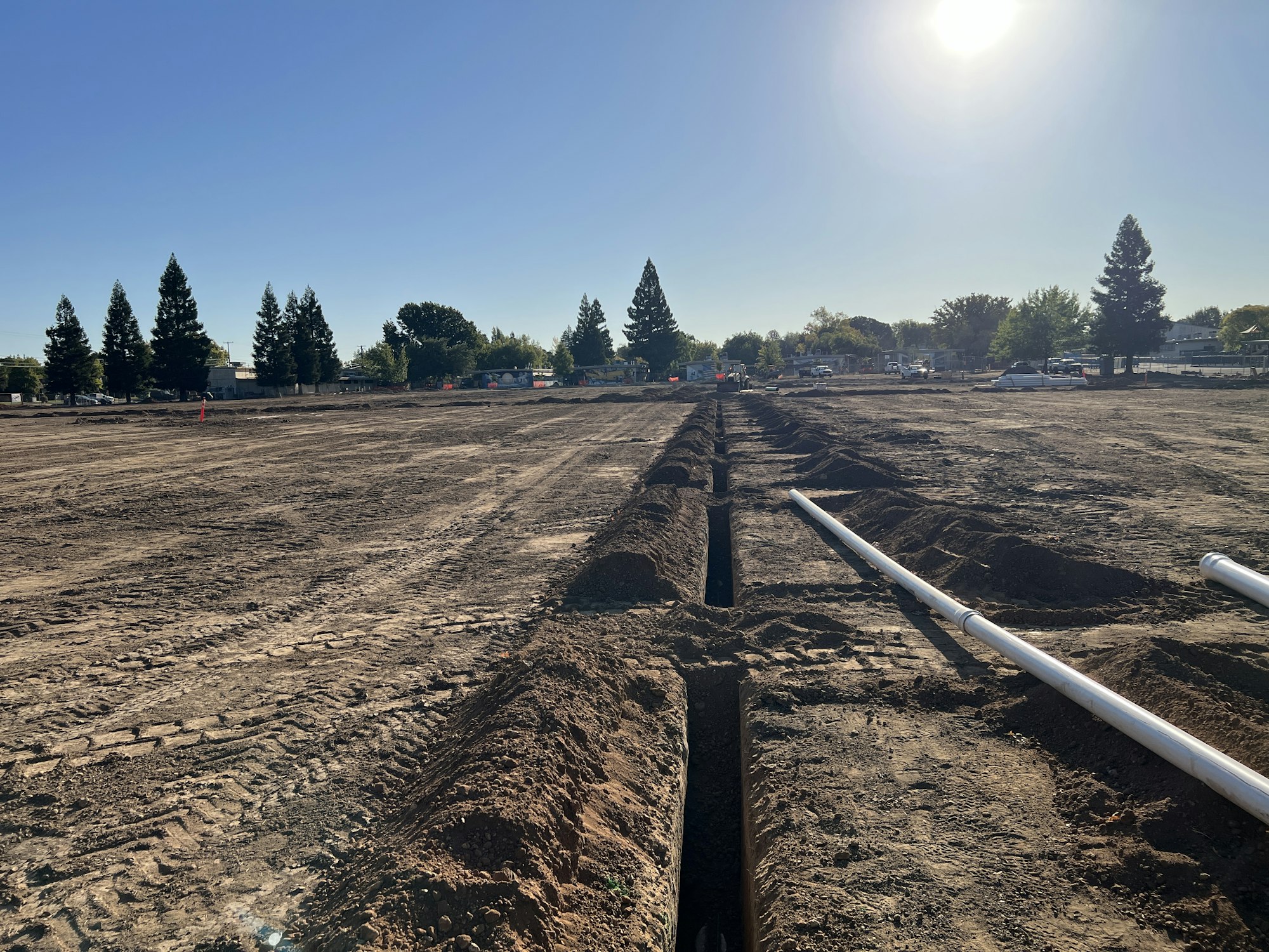Construction site with trenches and pipes, surrounded by trees under a clear sky.